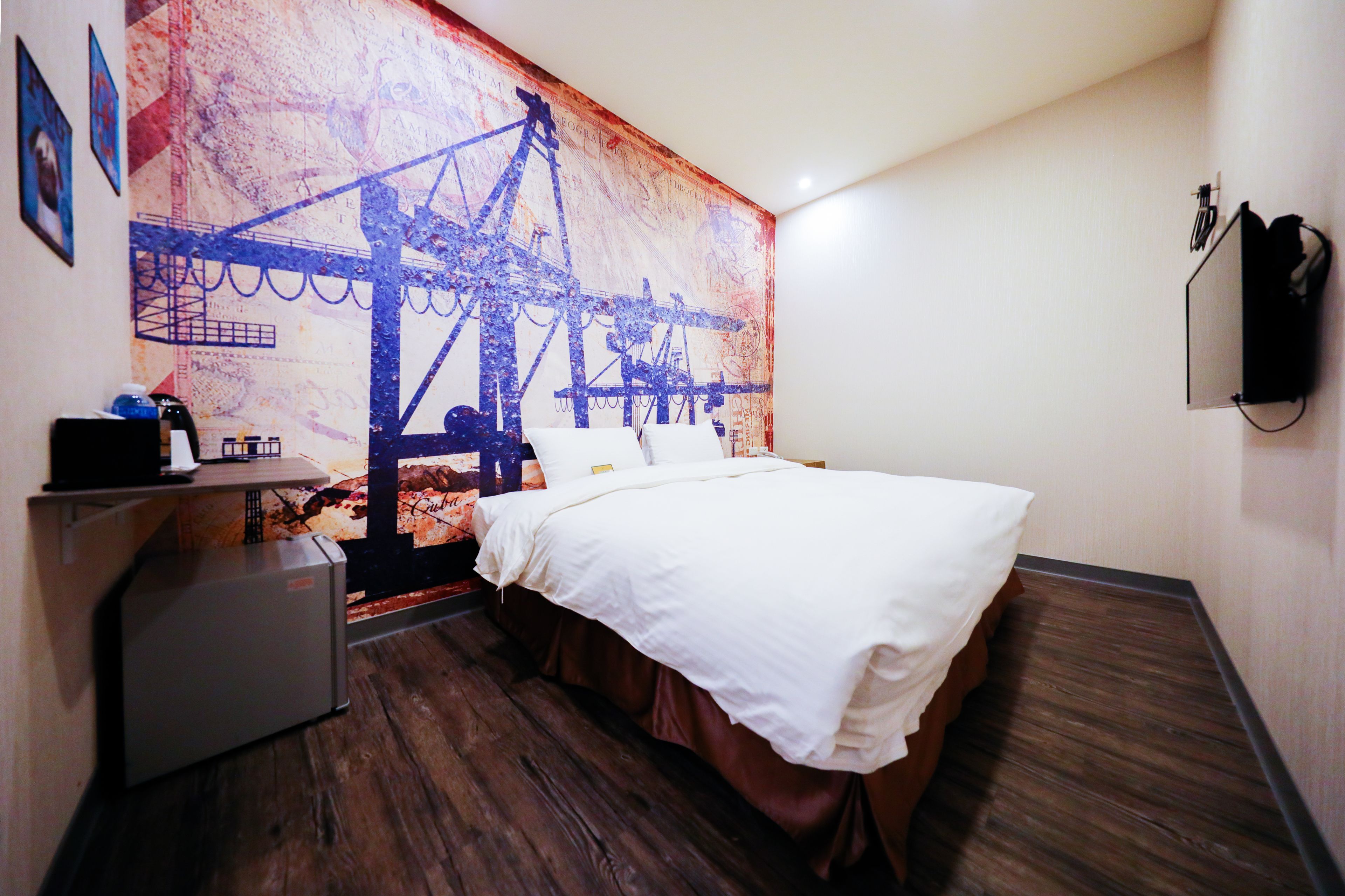 A hotel room with a bed, a desk, a refrigerator, and a wall with a large artwork.