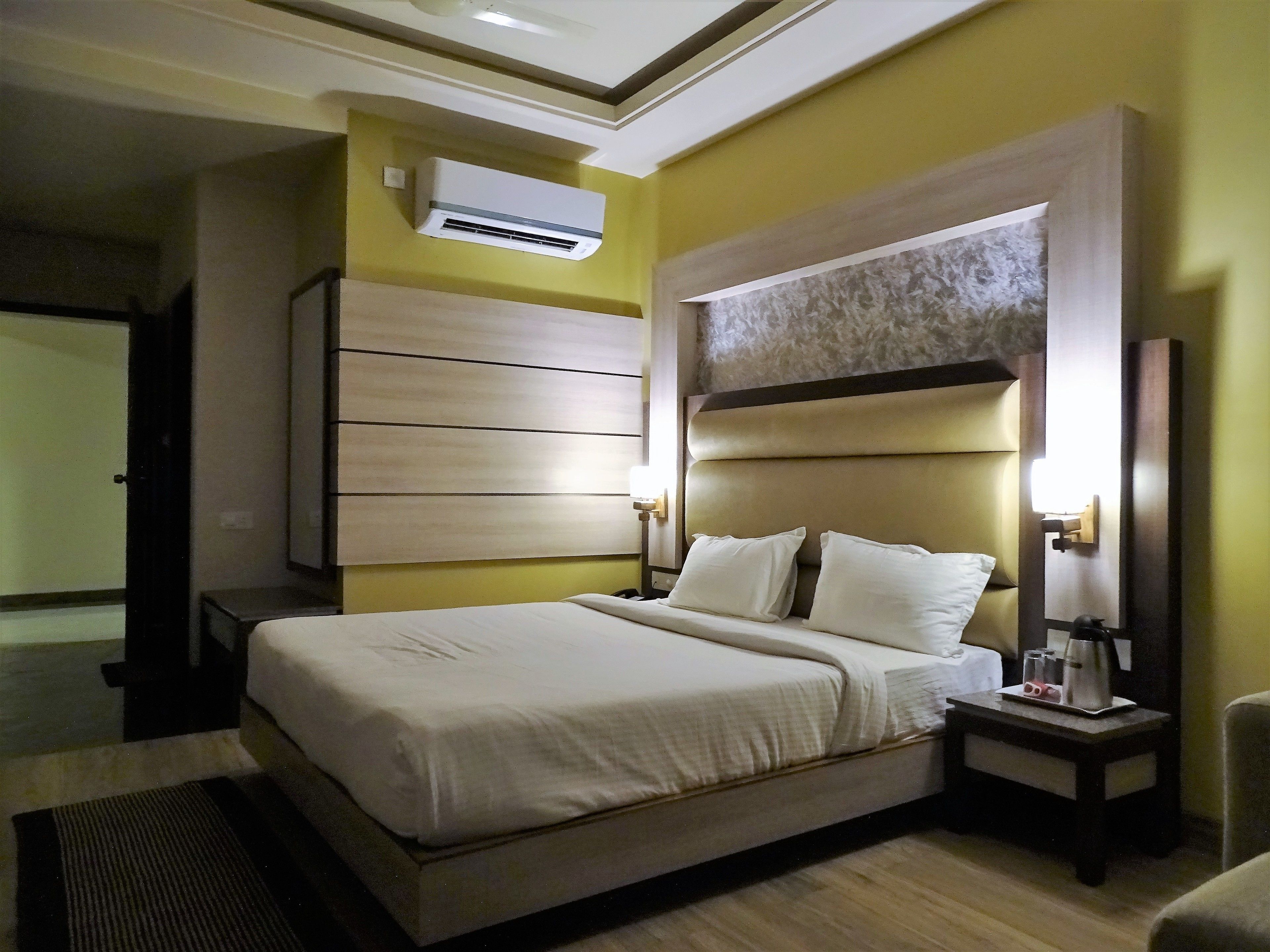A hotel room with a large bed, a bedside table with a lamp, an air conditioner, and a wall-mounted artwork.