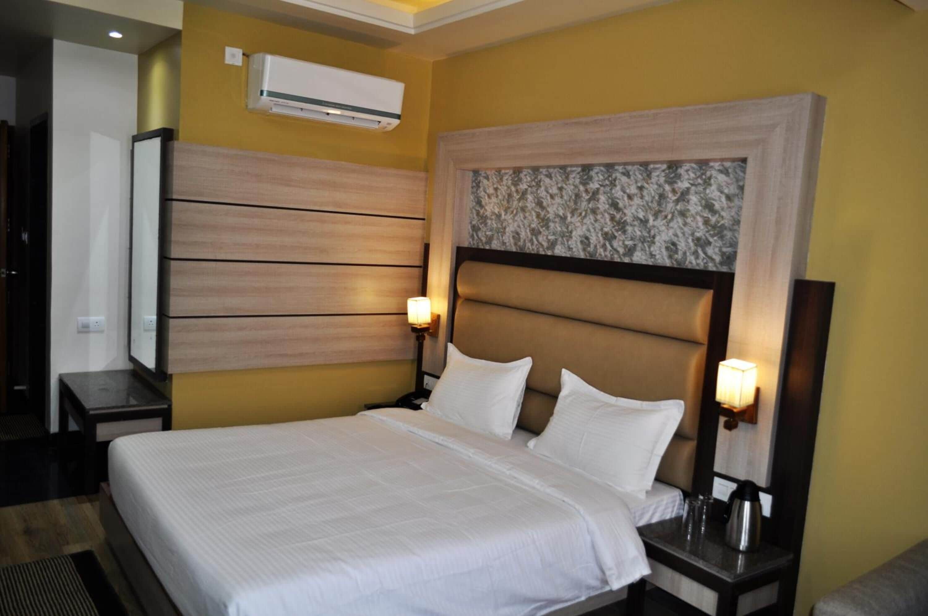 A hotel room with a large bed, two bedside tables, a wall-mounted air conditioner, and a decorative headboard.