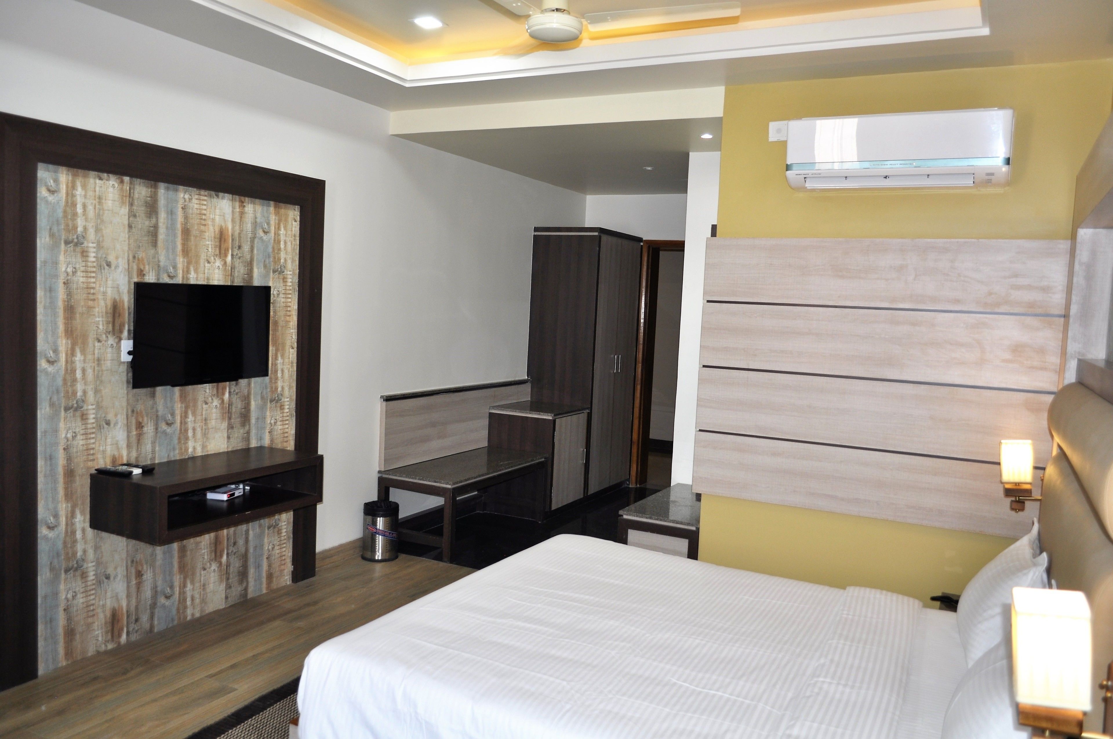 A hotel room with a bed, a desk, a wardrobe, a television mounted on a wooden wall, and an air conditioning unit on the wall.