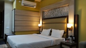 A hotel room with a large bed, two bedside lamps, an air conditioning unit, and a small table with a kettle and cups.