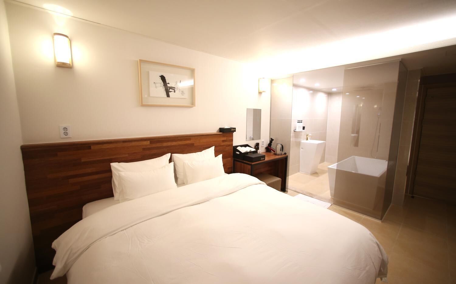 Una habitación de hotel con cabecera de madera, cama con ropa blanca, mesita de noche con lámpara, un cuadro enmarcado en la pared y un baño con mampara de vidrio.