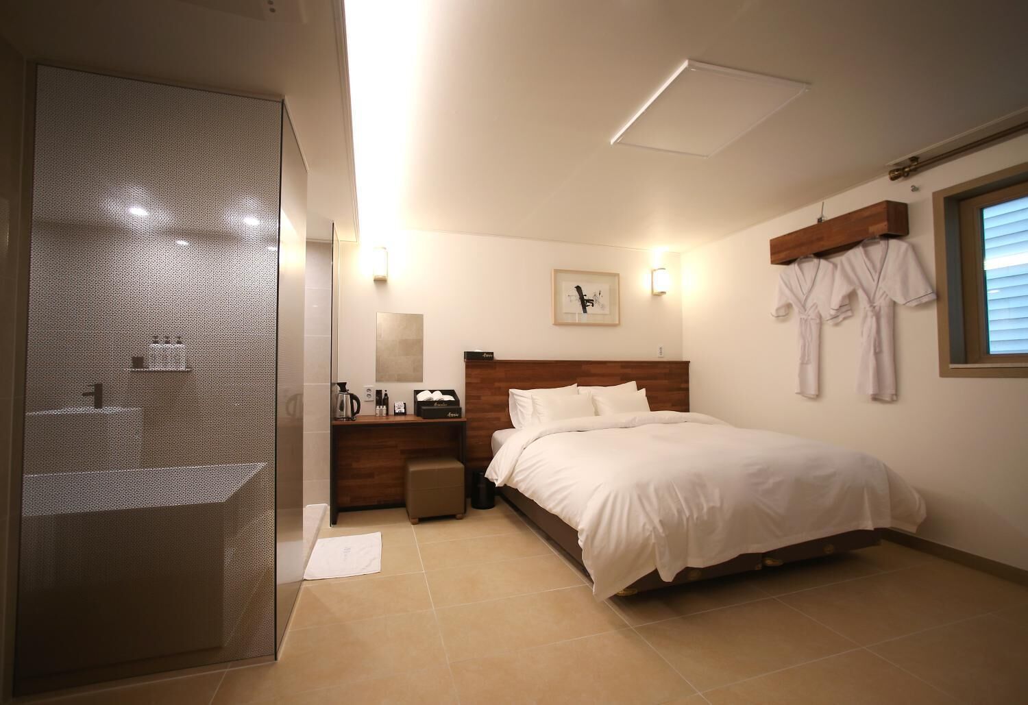 Une chambre d’hôtel comprenant un lit, une tête de lit en bois, une table de chevet, un miroir, une fenêtre avec des stores et un porte-peignoir.