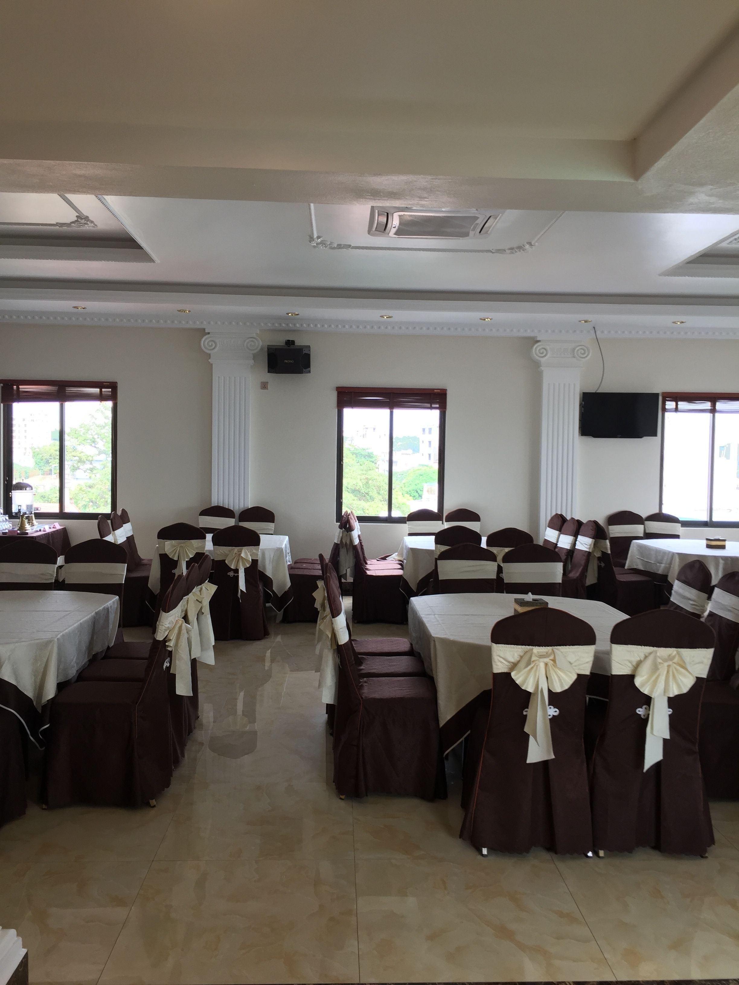 Un salón de banquetes con varias mesas y sillas, ventanas amplias que dan a zonas verdes y un televisor de pantalla plana montado en la pared.