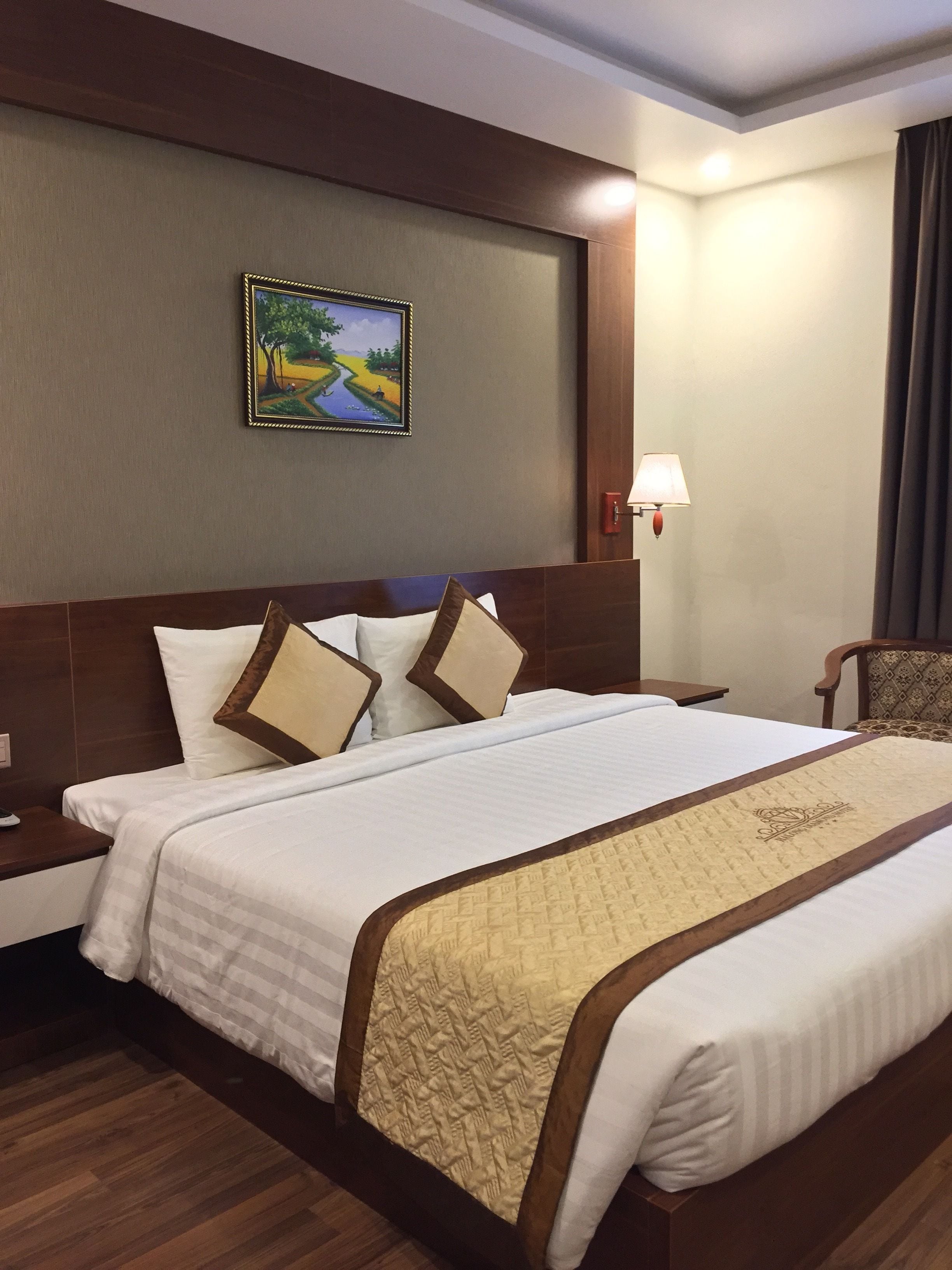 A hotel room with a large bed, a nightstand, a lamp, a chair, and a painting on the wall.