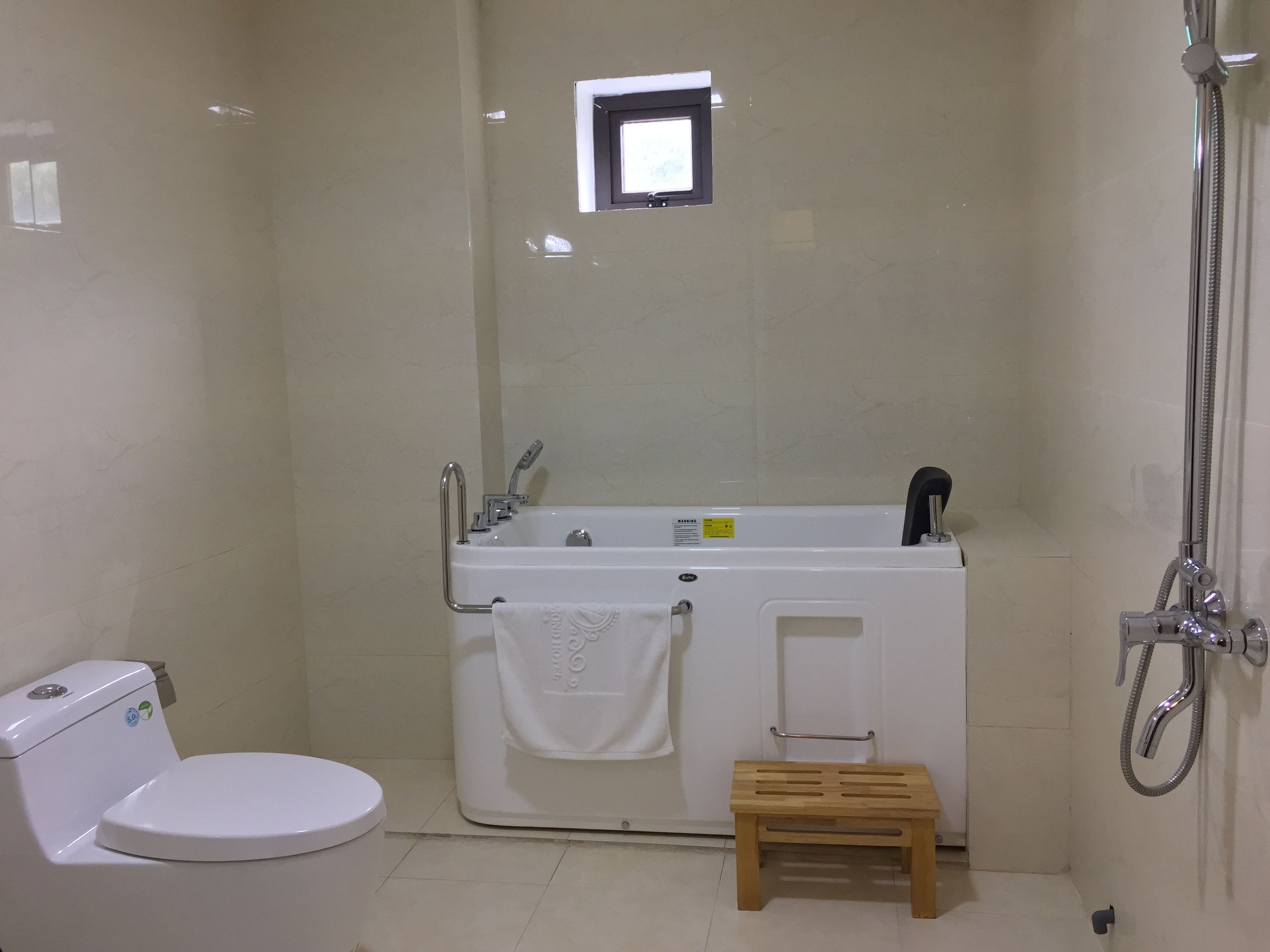 A small, clean bathroom with a white toilet, a bathtub with a shower head, and a small wooden stool.