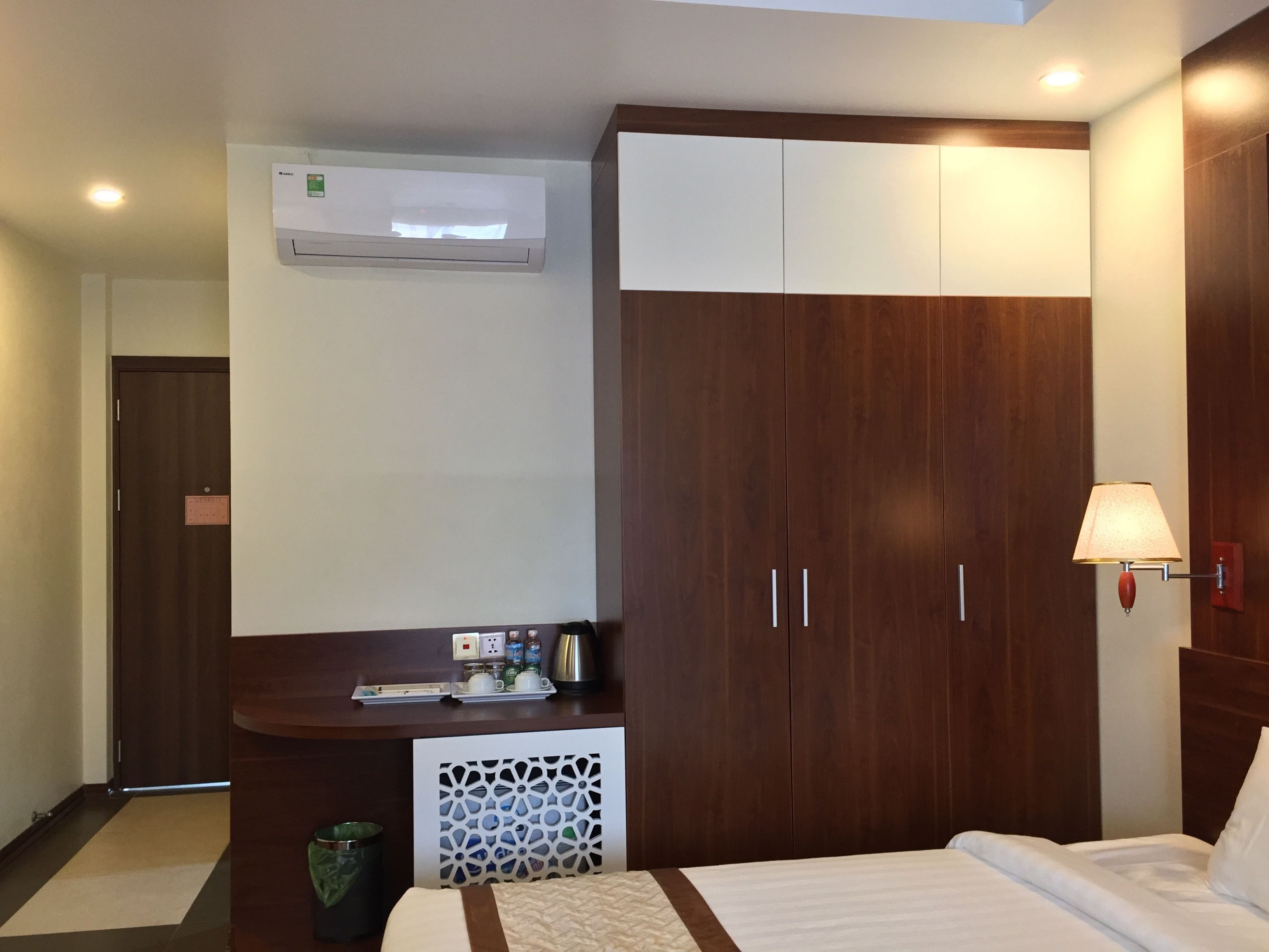 A hotel room with a bed, a wooden wardrobe, a small table with toiletries, an air conditioning unit, and a wall-mounted lamp.