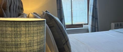 A neatly made bed with white linens, a wooden nightstand with a lamp, and a window with blue curtains.