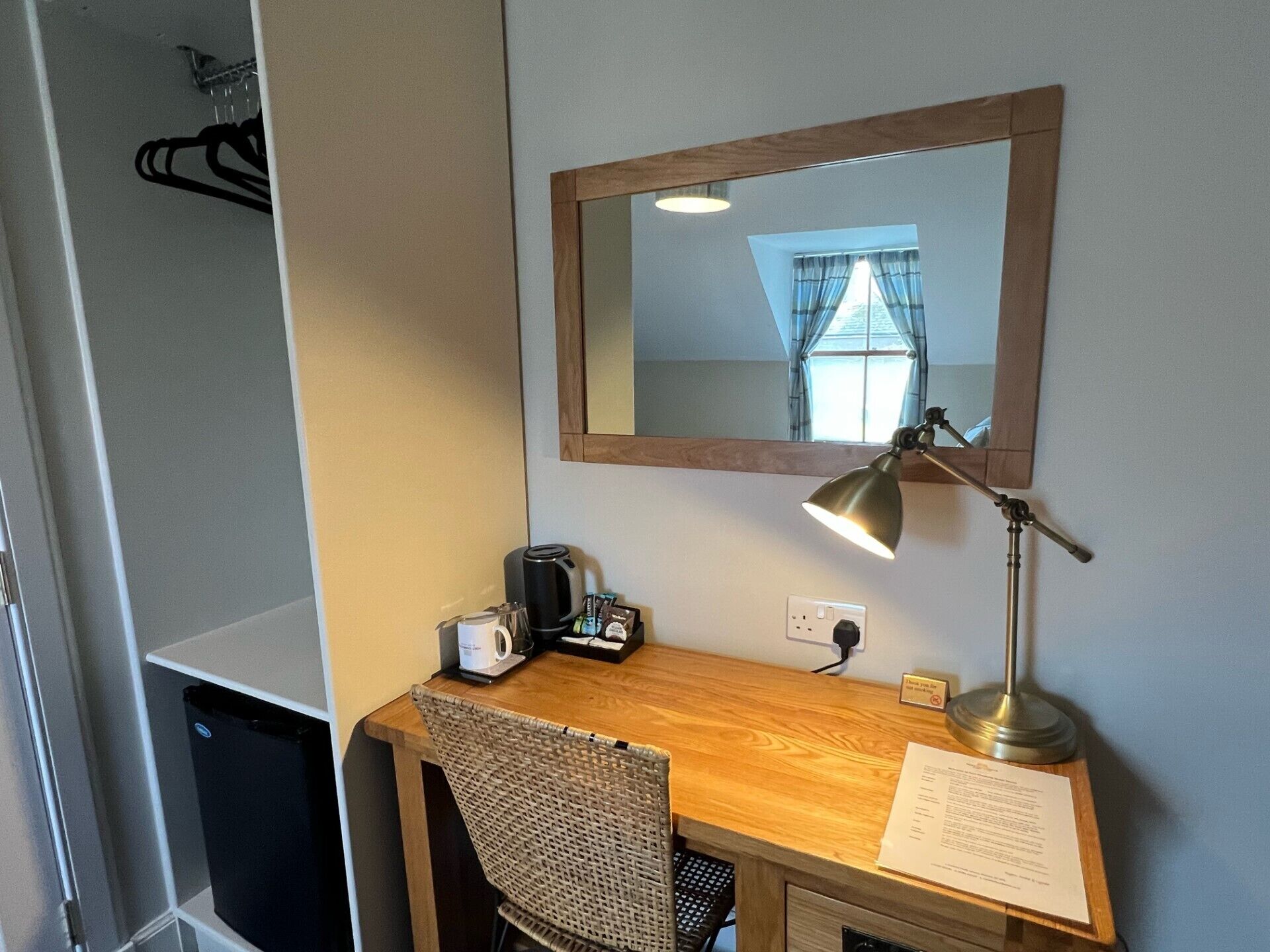 Ett hotellrum med ett skrivbord i trä, en stol, en spegel, en bordslampa, en kaffebryggare och en papperskorg.