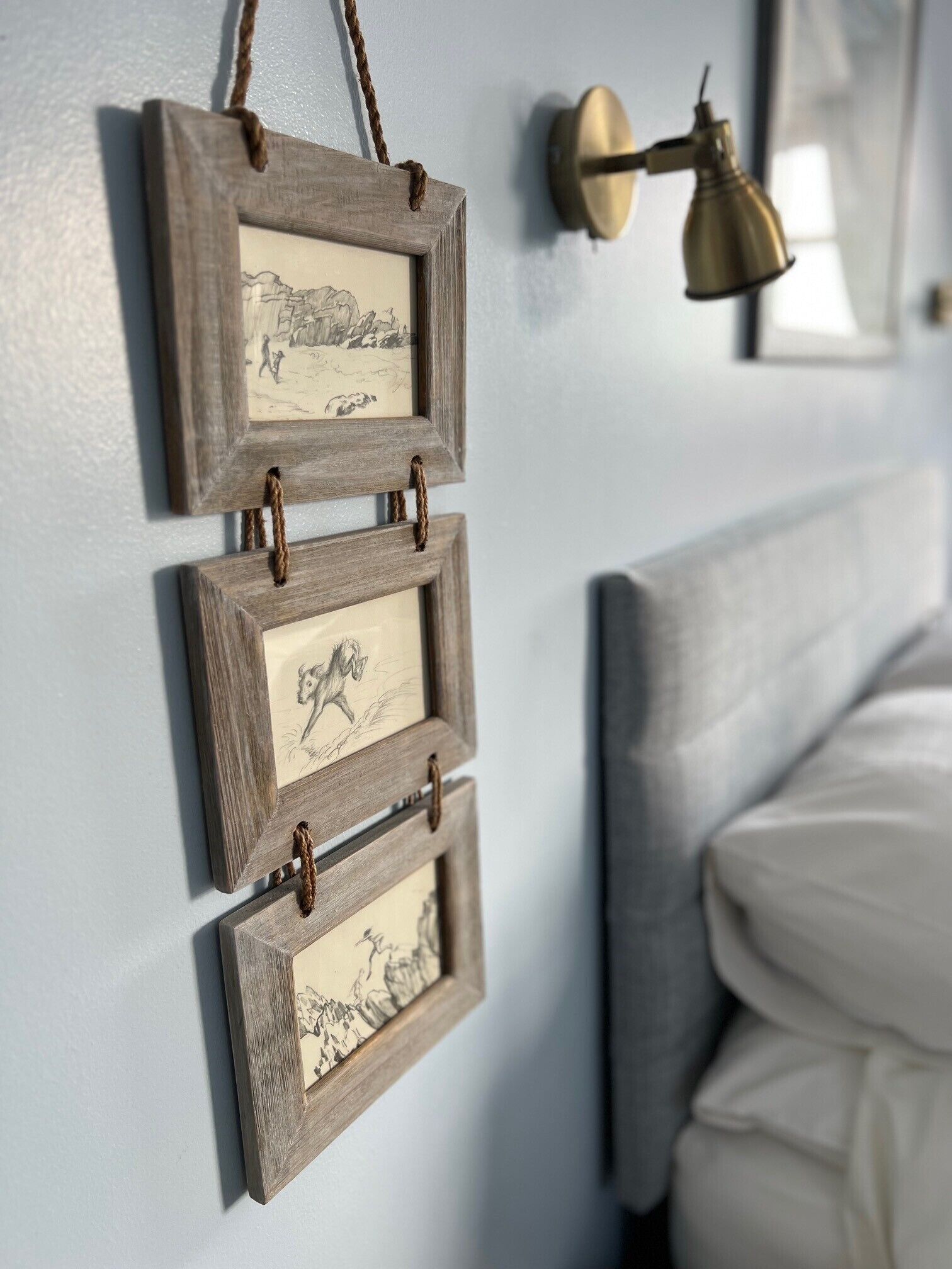A wall with three wooden picture frames hanging vertically, each containing a black and white drawing. A brass wall sconce is mounted above the frames.