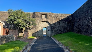 Uma trilha de pedra leva a um edifício com porta azul, cercado por muros de pedra e vegetação.