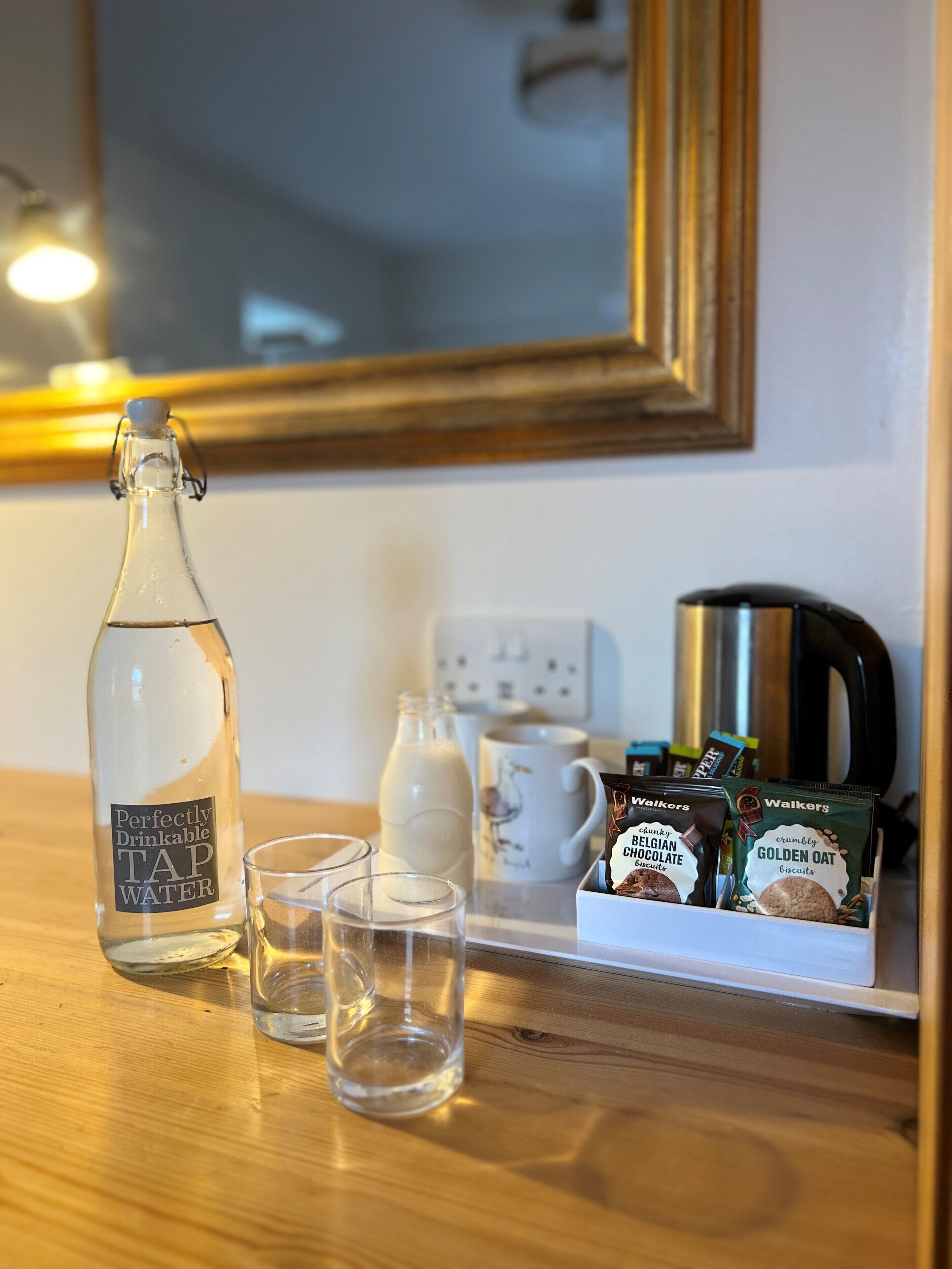 A kitchen counter with a bottle of tap water, two glasses, a milk jug, a coffee maker, and a box of biscuits.