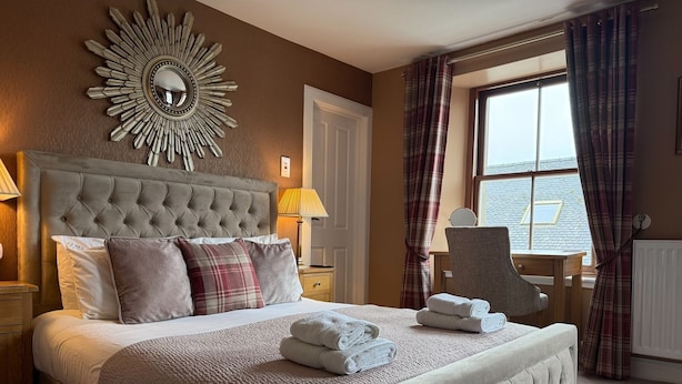 A bedroom with a tufted headboard, a large mirror, a bed with pillows, a bedside table with a lamp, and a window with curtains. - Fort Charlotte Guesthouse (Lerwick)