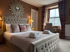 A bedroom with a tufted headboard, a large mirror, a bed with pillows, a bedside table with a lamp, and a window with curtains. - Fort Charlotte Guesthouse (Lerwick)