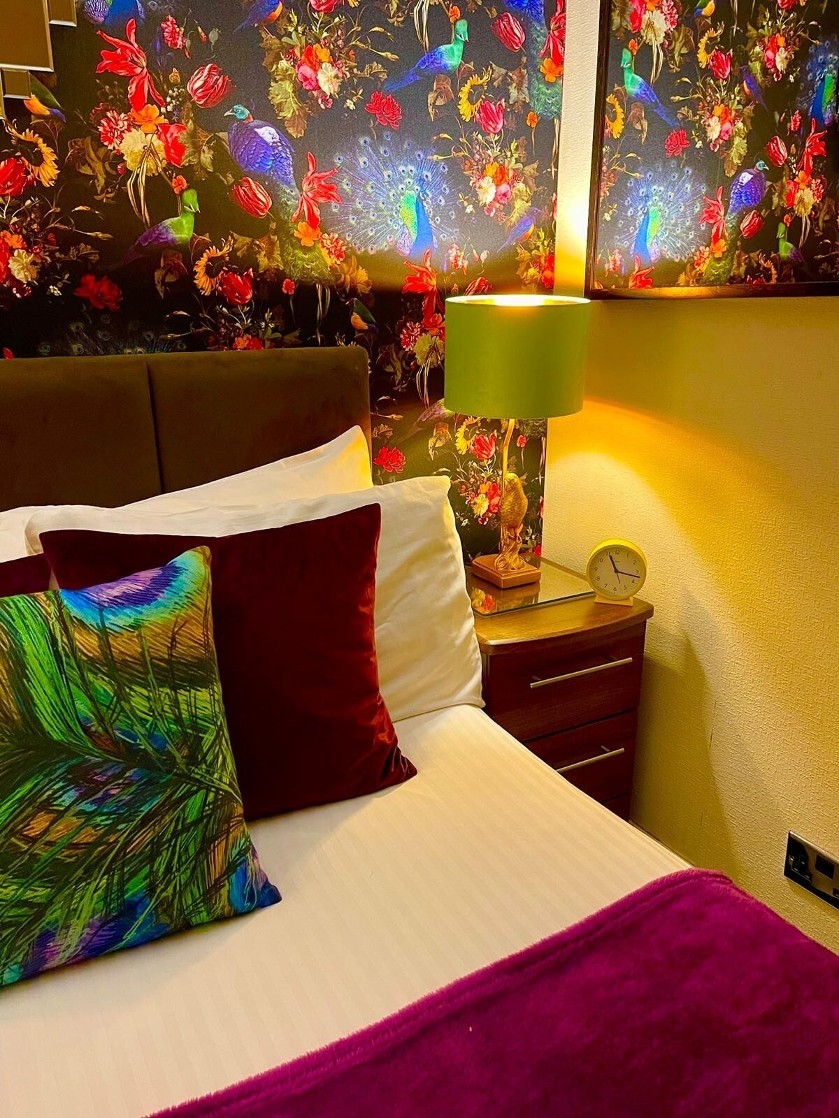 A bed with a colorful pillow, a green lamp, and a clock on a bedside table.