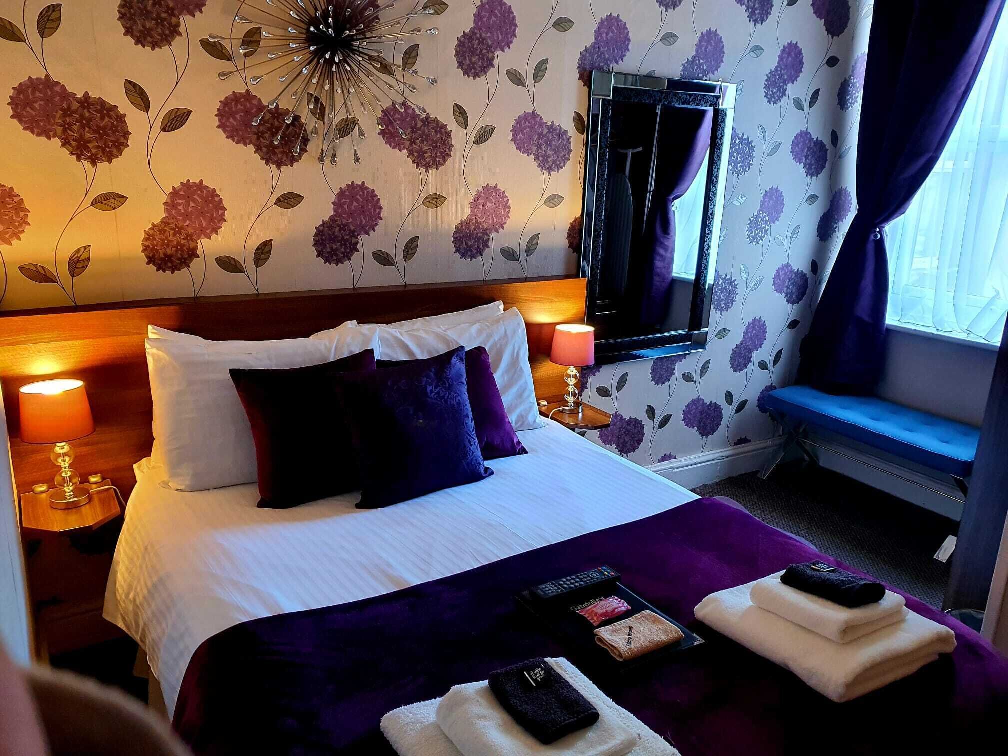 A hotel room with a bed, a TV, a window with curtains, a bedside table with a lamp, and a wall with floral wallpaper.