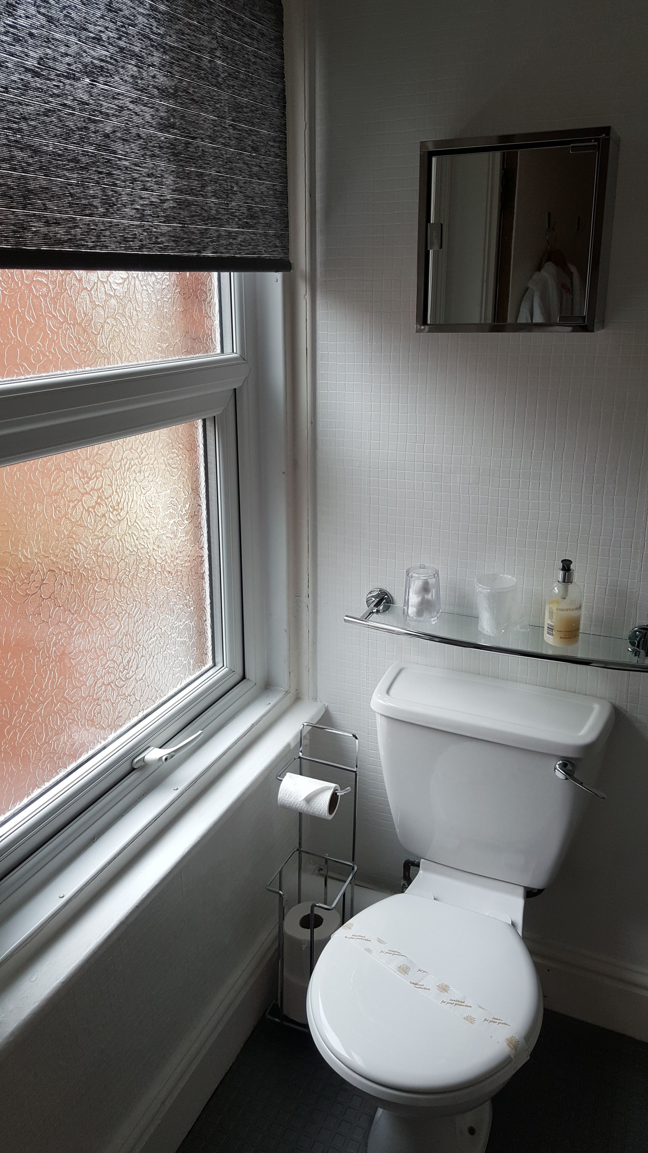 A bathroom with a toilet, a glass shelf with toiletries, a window with a frosted glass, and a mirror on the wall.