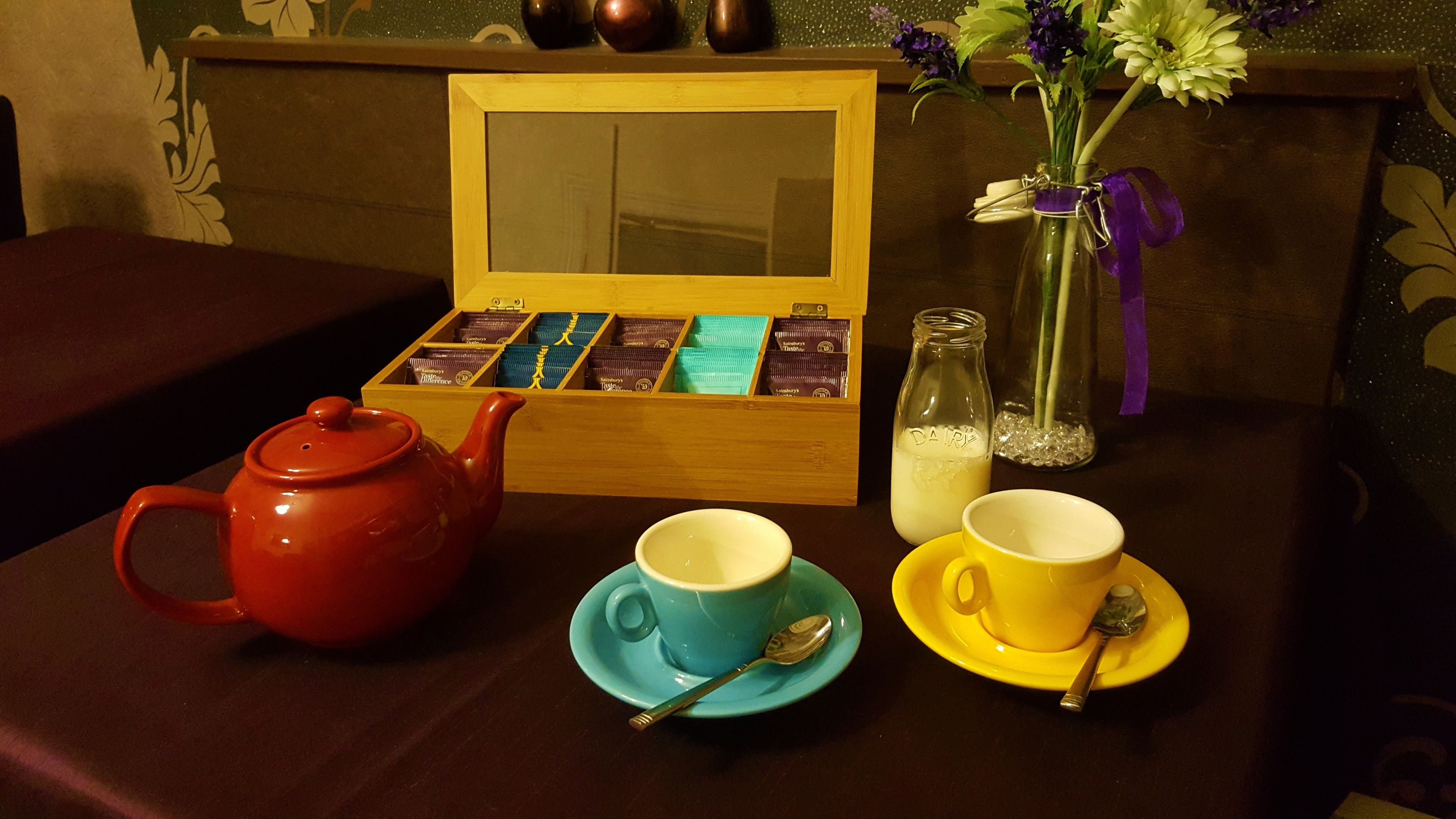 A table with a red teapot, two cups with saucers, a spoon, a glass bottle, and a box of chocolates.
