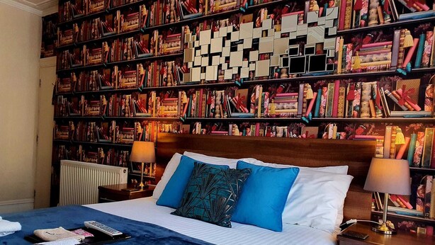 Une chambre d’hôtel avec une grande bibliothèque remplie de livres, un lit avec une literie bleue et blanche, et deux tables de chevet avec des lampes.