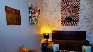 Un salon chaleureux avec un canapé marron, deux coussins bleus, une table en bois sur laquelle est posé un jouet en forme de canard, et des œuvres d’art murales comprenant une enseigne « Pêche au canard » et une affiche de cirque.