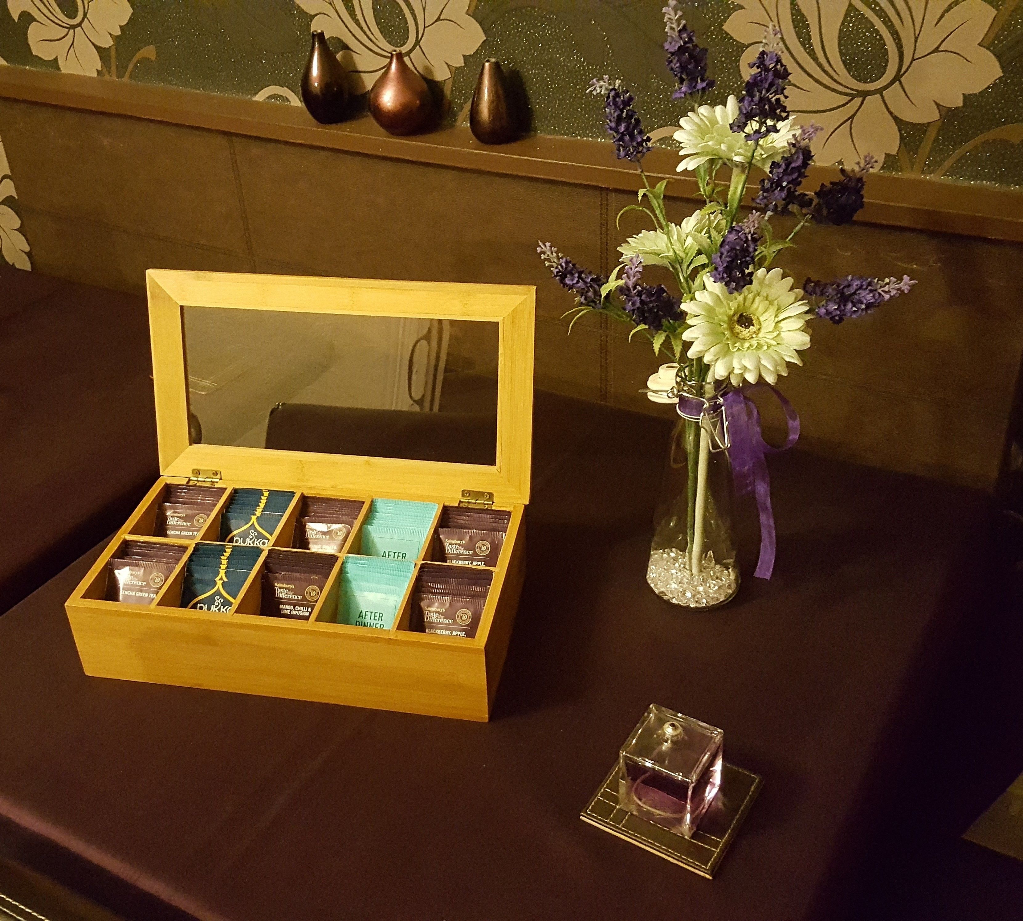 A wooden box with compartments containing various tea products, a vase with flowers, and a small glass container on a table.