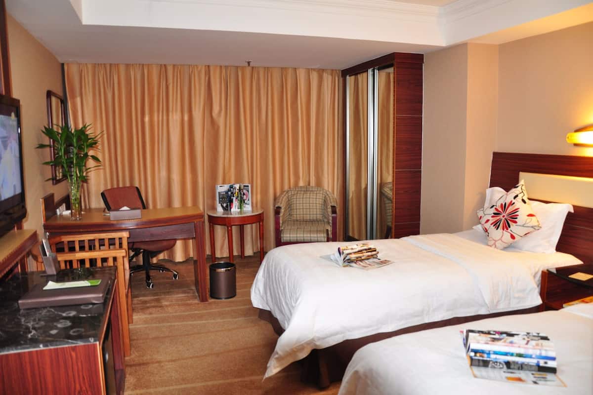 A hotel room with two beds, a desk with a laptop, a chair, a small table with a plant, and a large window with curtains.