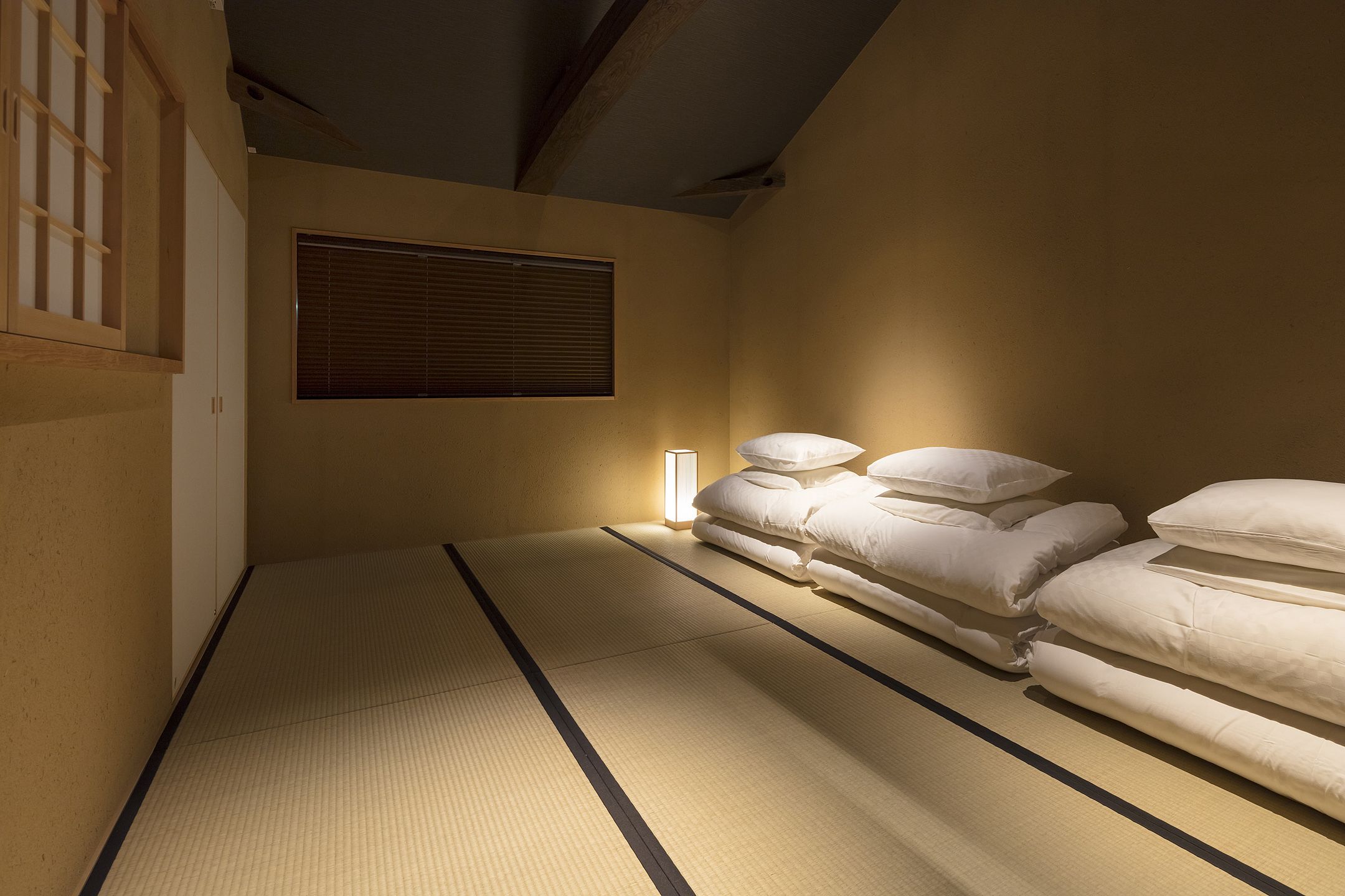 Una habitación con suelo de tatami, una cama, un ventanal con persianas y una mesita de noche con lámpara.