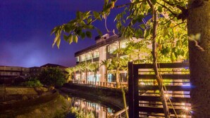 Un bâtiment moderne à plusieurs étages, avec une étendue d’eau réfléchissante devant et un arbre aux feuilles vertes au premier plan.