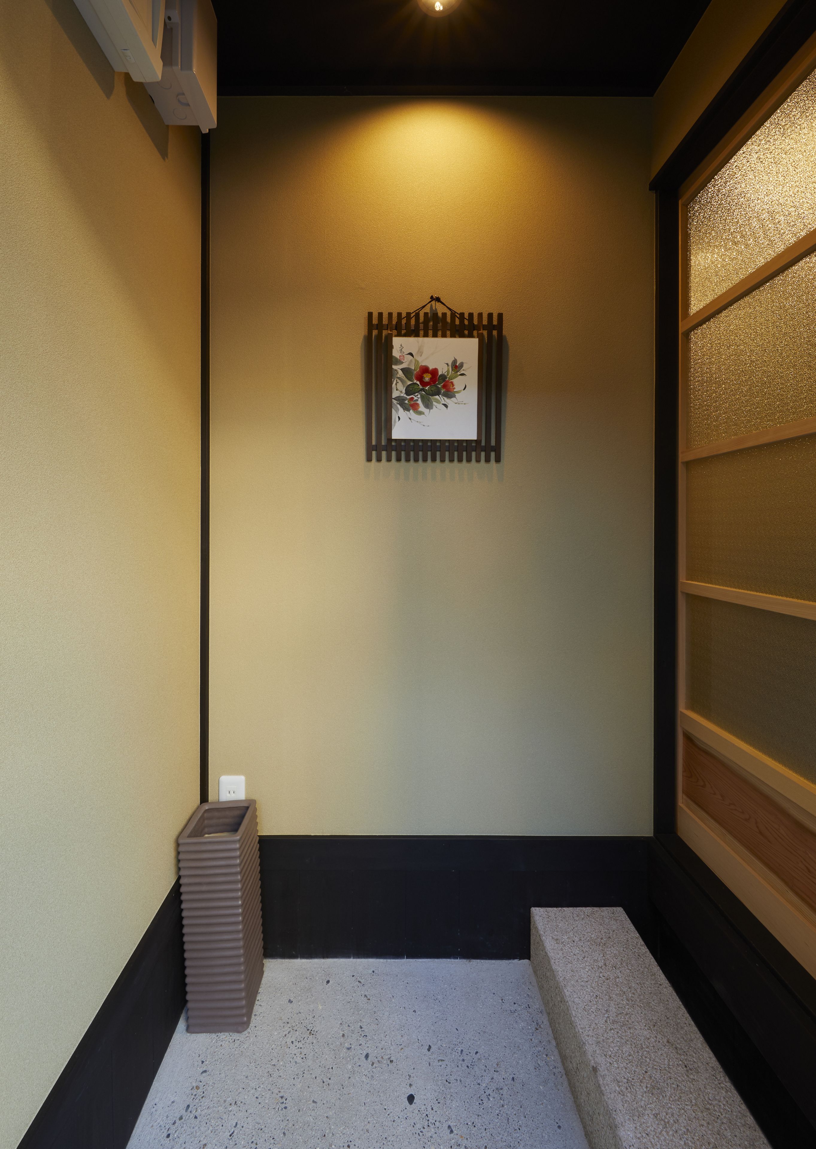 A small, dimly lit room with a single framed artwork on the wall, a trash bin, and a textured glass partition.