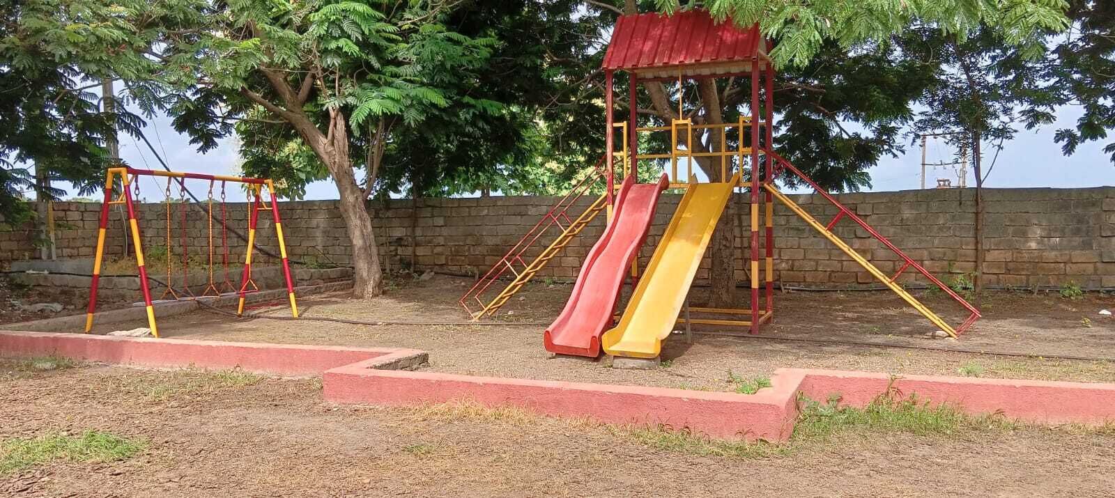 Un parque infantil con columpios y toboganes, rodeado de árboles y un muro de ladrillo.