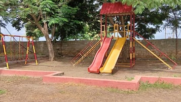 Un parque infantil con columpios y toboganes, rodeado de árboles y un muro de ladrillo.