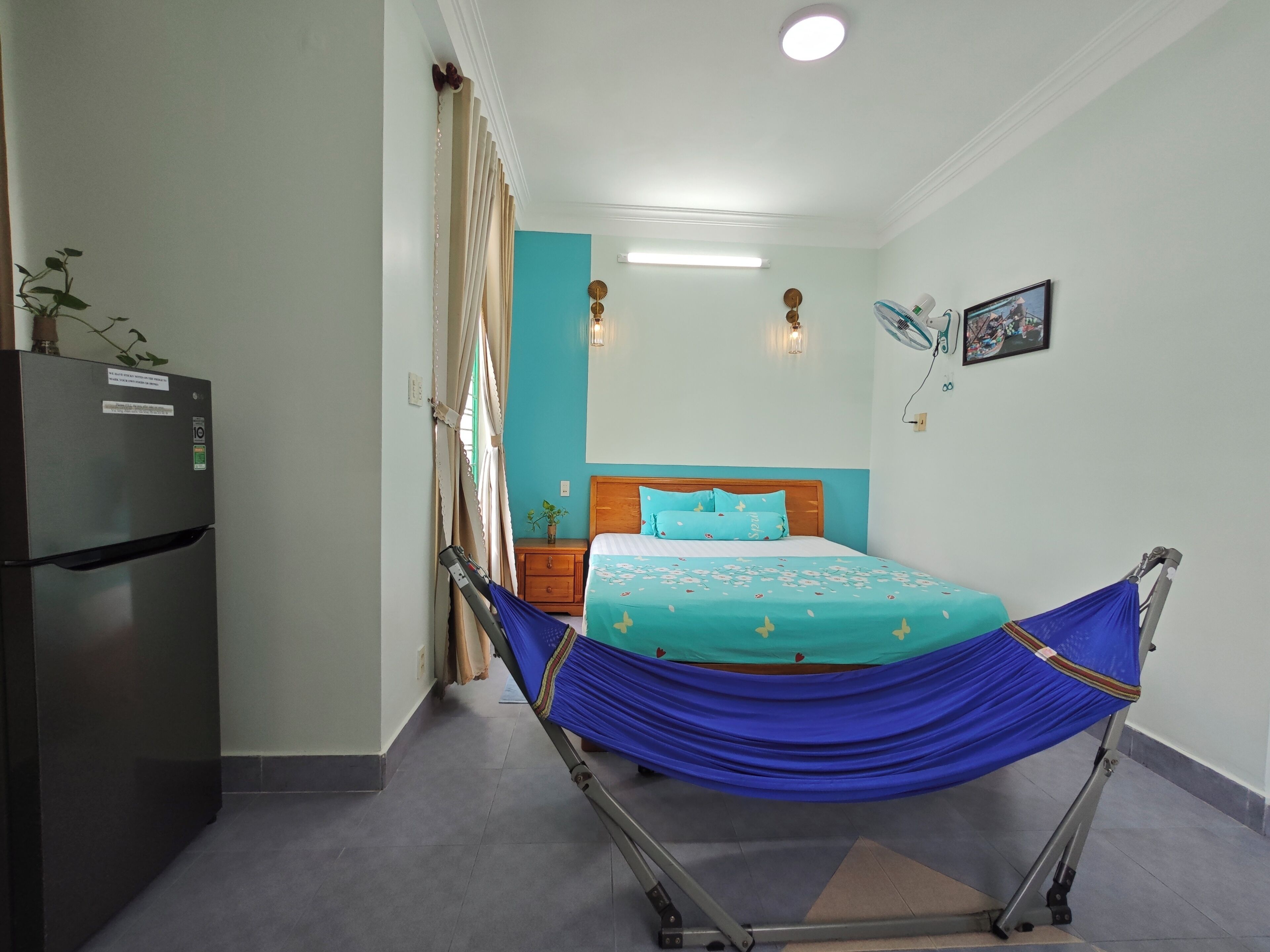 Ein Schlafzimmer mit Bett, Nachttisch, Kühlschrank, einem Fenster mit Vorhängen und einem Wandventilator.