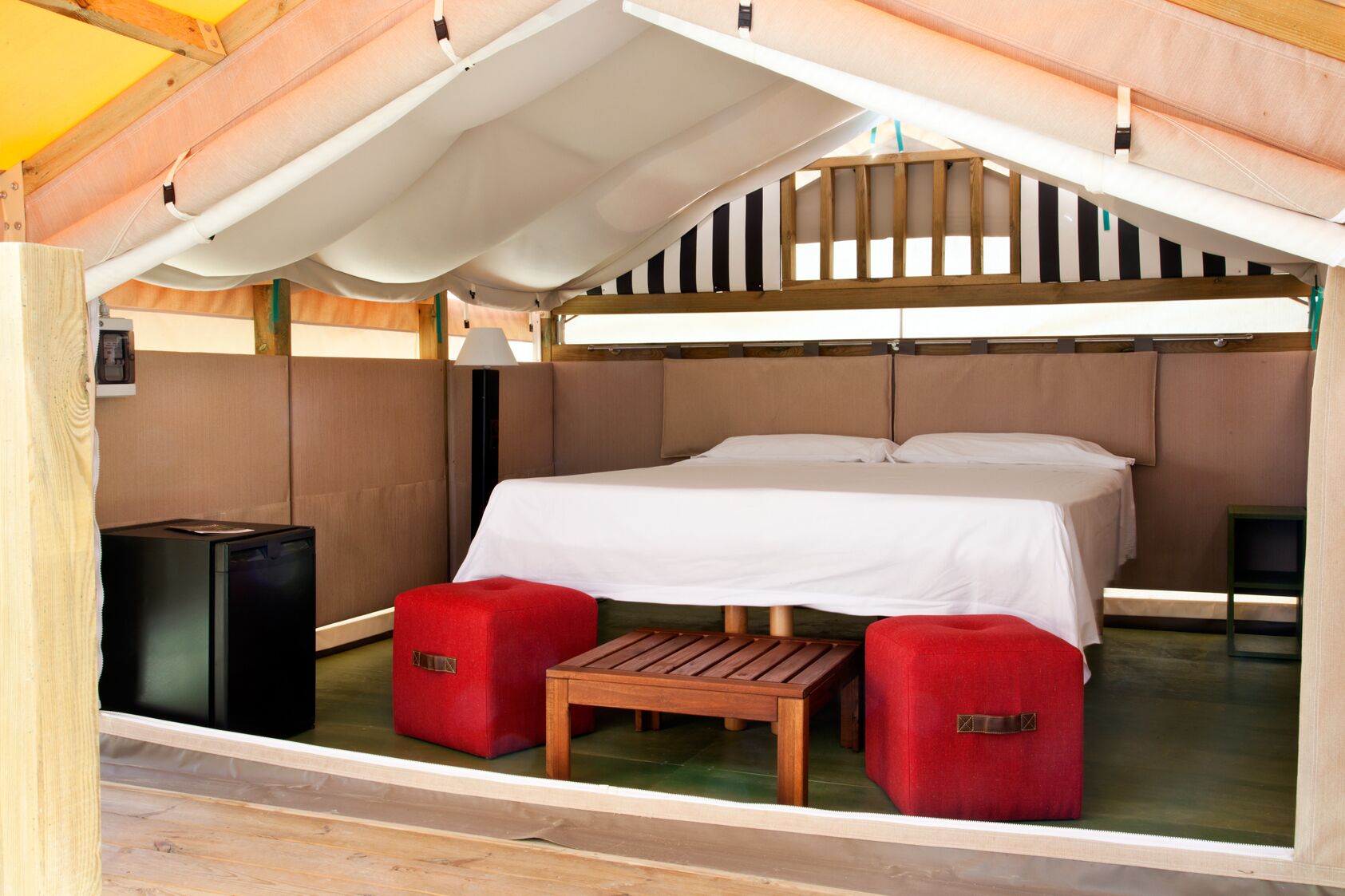 Eine gemütliche Einzimmerhütte mit Bett, einem kleinen Tisch, zwei roten Hocker und einem schwarzen Lautsprecher.