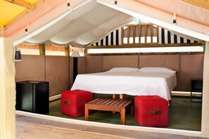 Una cabaña pequeña y de una sola habitación, con cama, una mesita, dos otomanas rojas y un altavoz negro.