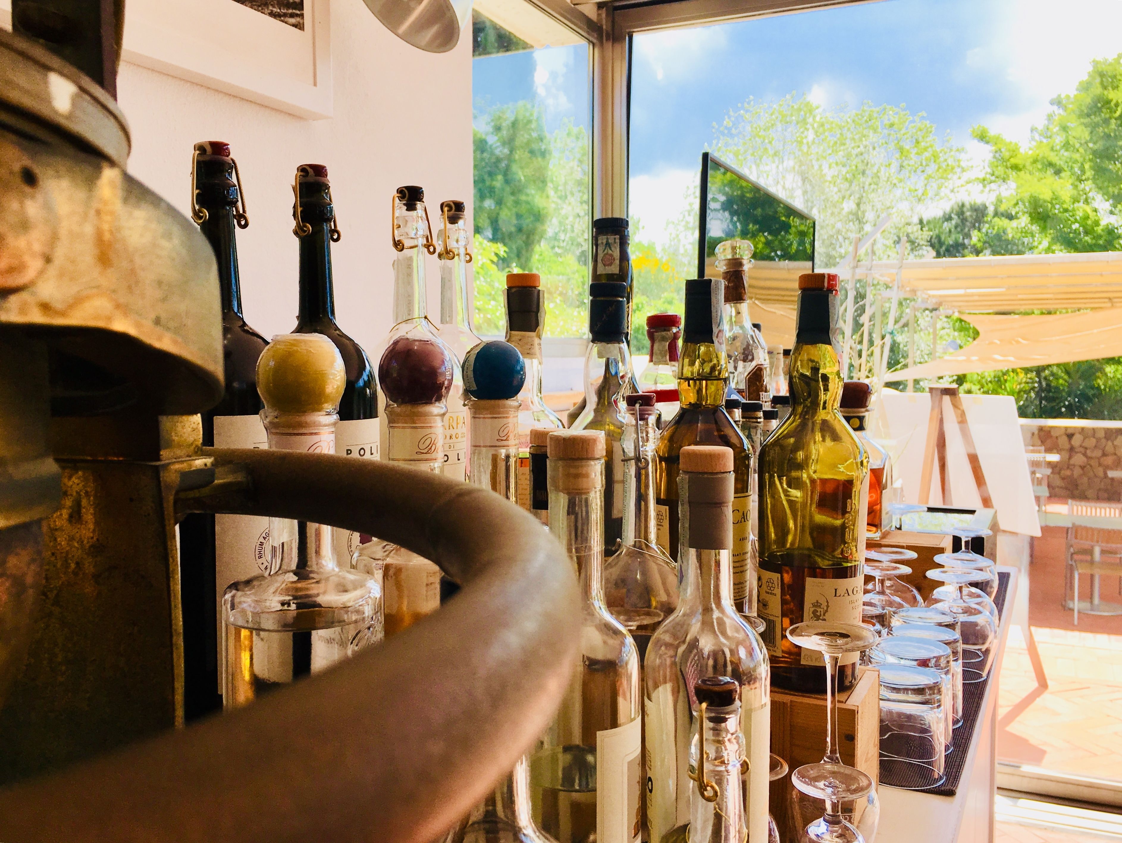 Zona de bar con diversas botellas y vasos, un barril de madera grande y vistas a árboles a través de una ventana.