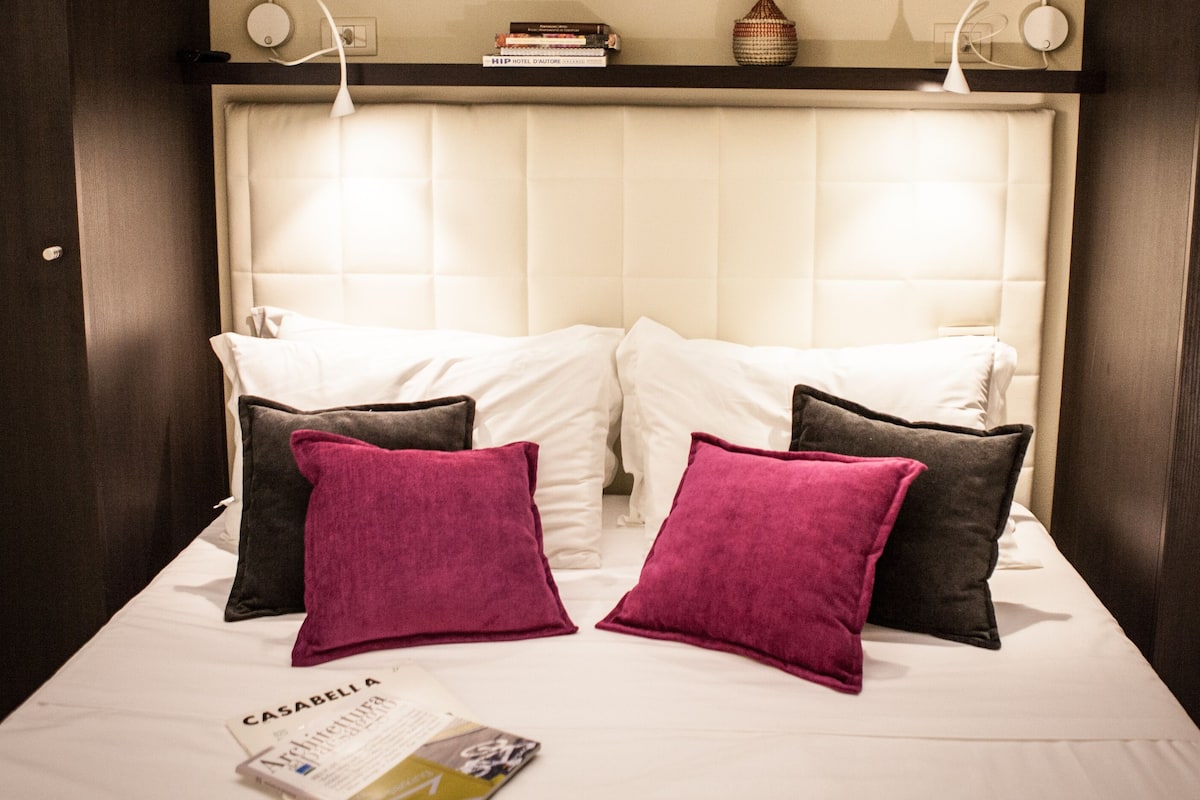 A hotel room with a bed, two pillows, a magazine, and a shelf with books and a vase.