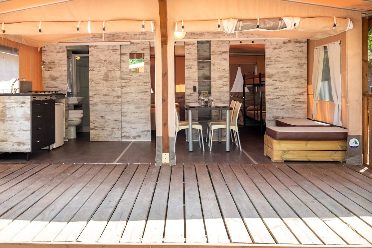 A covered outdoor area with a wooden deck, a dining table with chairs, a sofa, and a small kitchen area with a sink and cabinets.