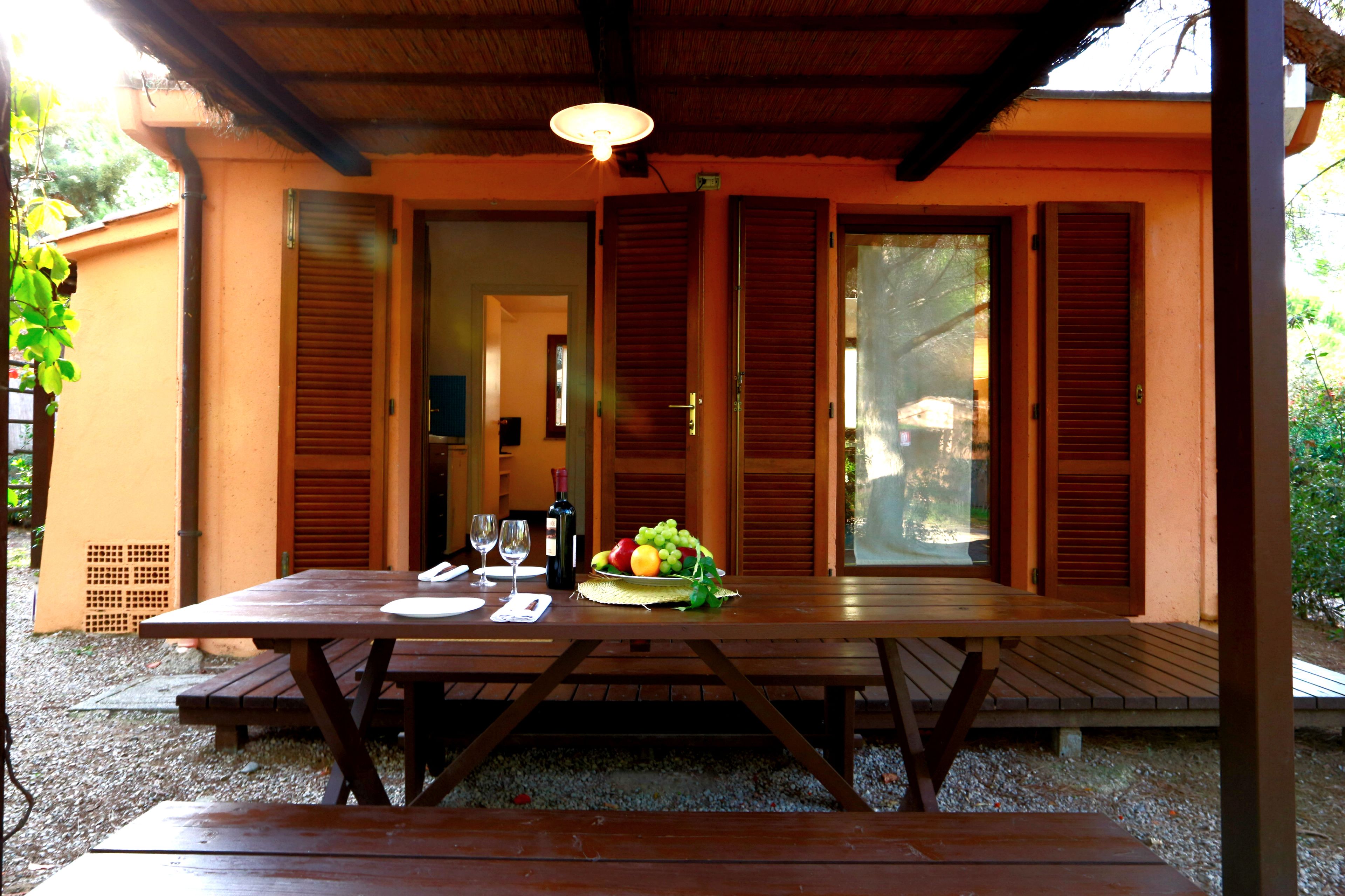 A wooden picnic table with a fruit basket and wine glasses set up under a covered patio.
