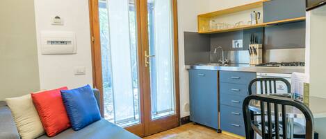 A compact kitchen with a sink, stove, and blue cabinets. A gray sofa with red and blue cushions is in the foreground.
