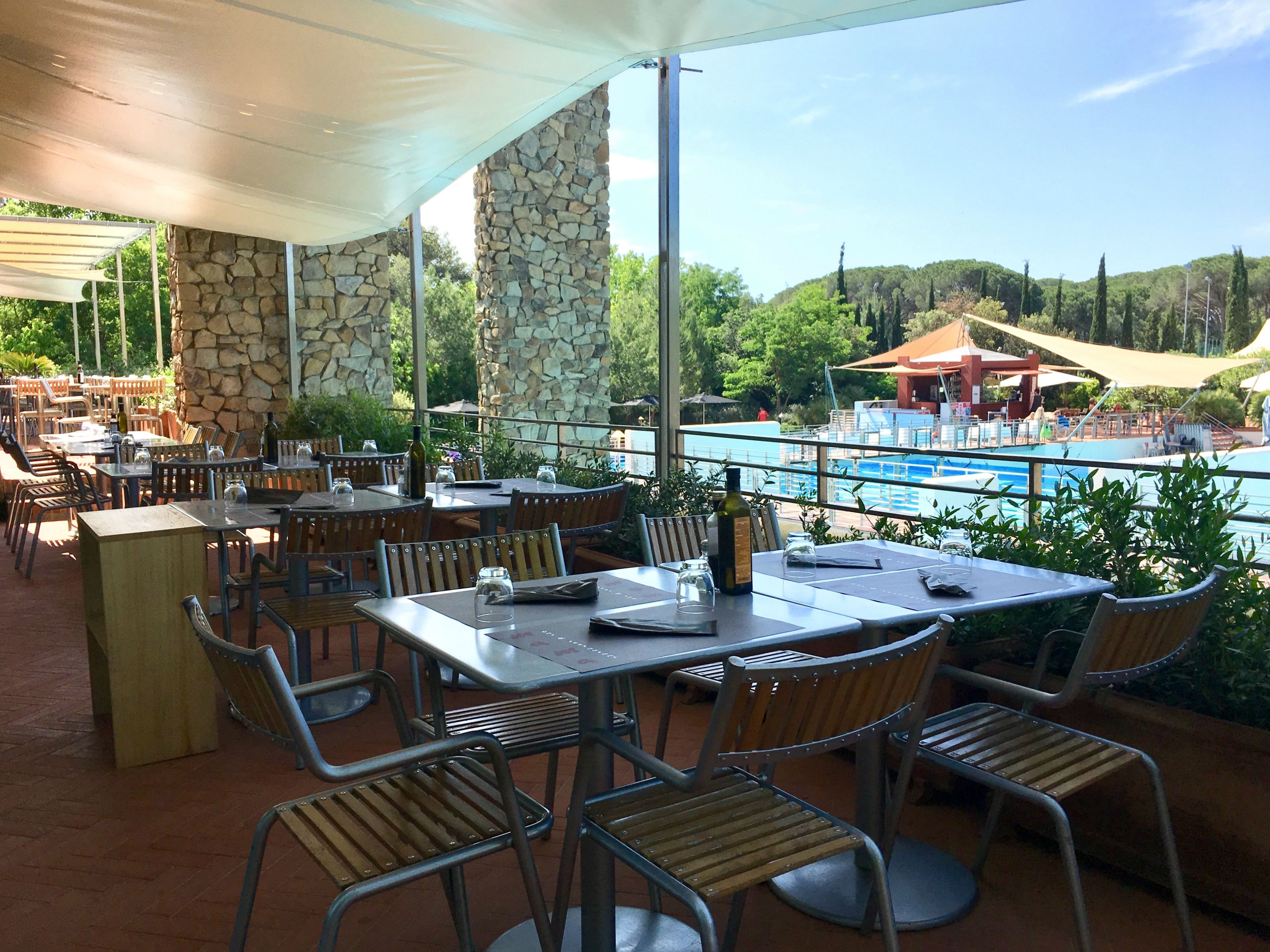 An outdoor dining area with tables and chairs, a stone wall, and a view of a pool and greenery.