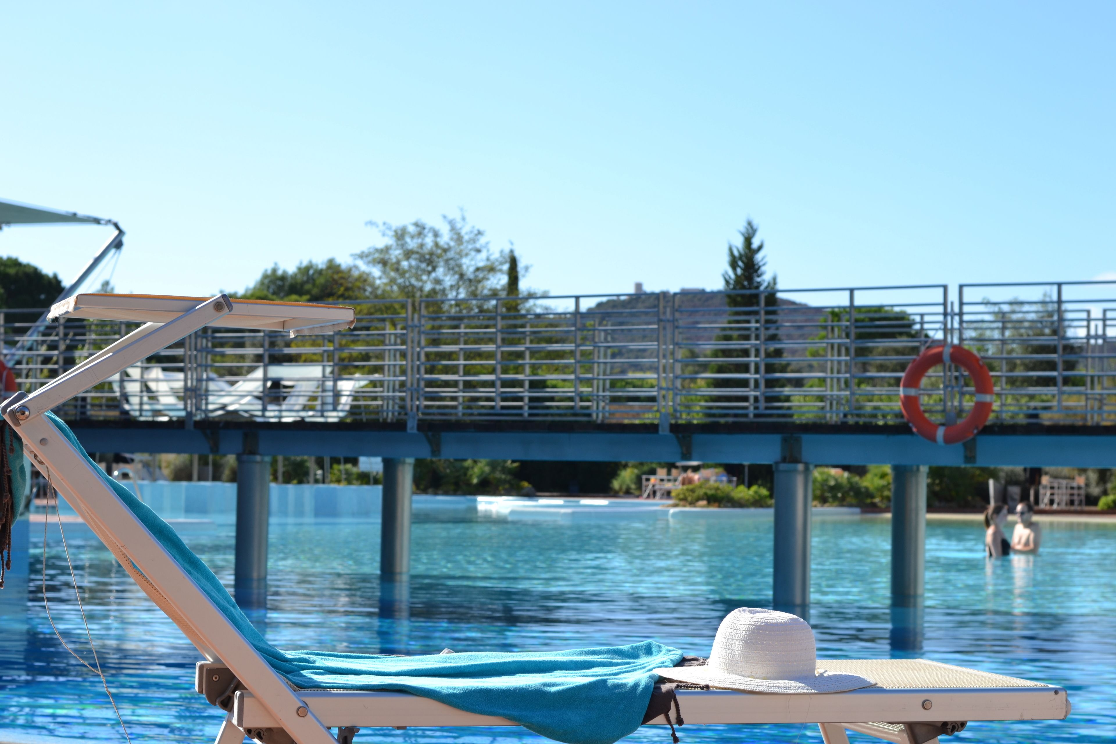 Una silla de descanso junto a la piscina, con una toalla y un sombrero, un flotador y un puente sobre el agua.