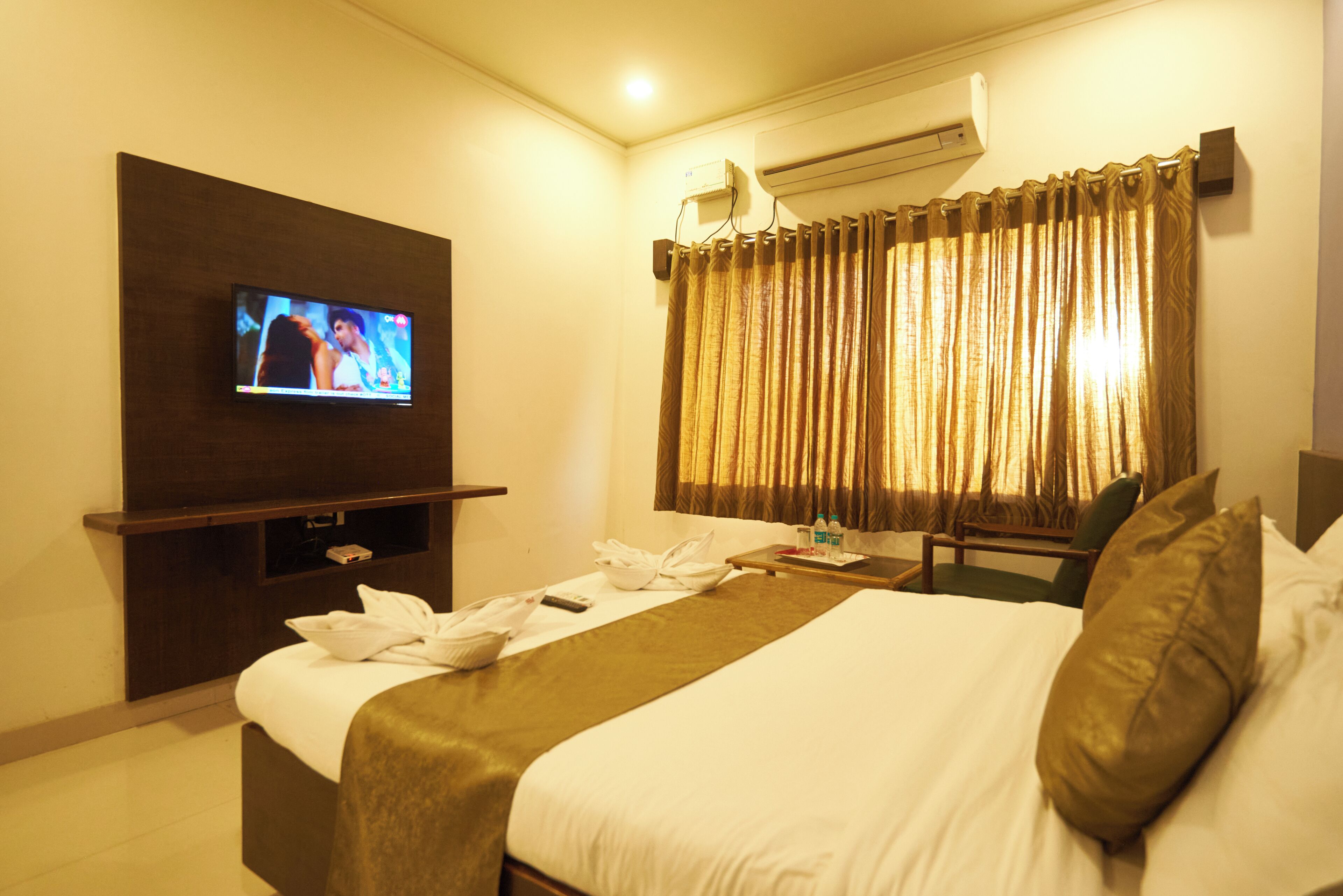 Una habitación de hotel con cama, televisor montado en la pared, ventana con cortinas, una silla y una mesita de noche con una botella y vasos.