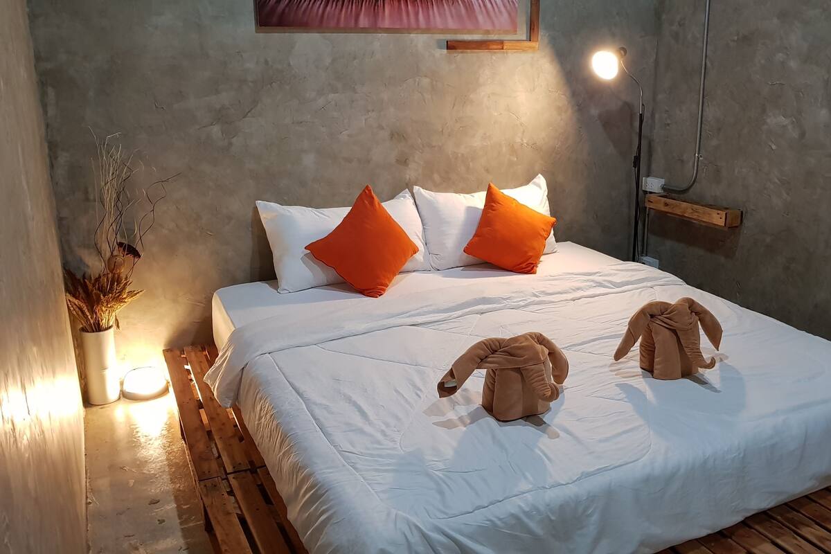 Cama bem arrumada com almofadas brancas e laranja, piso de madeira e luminária fixada na parede.