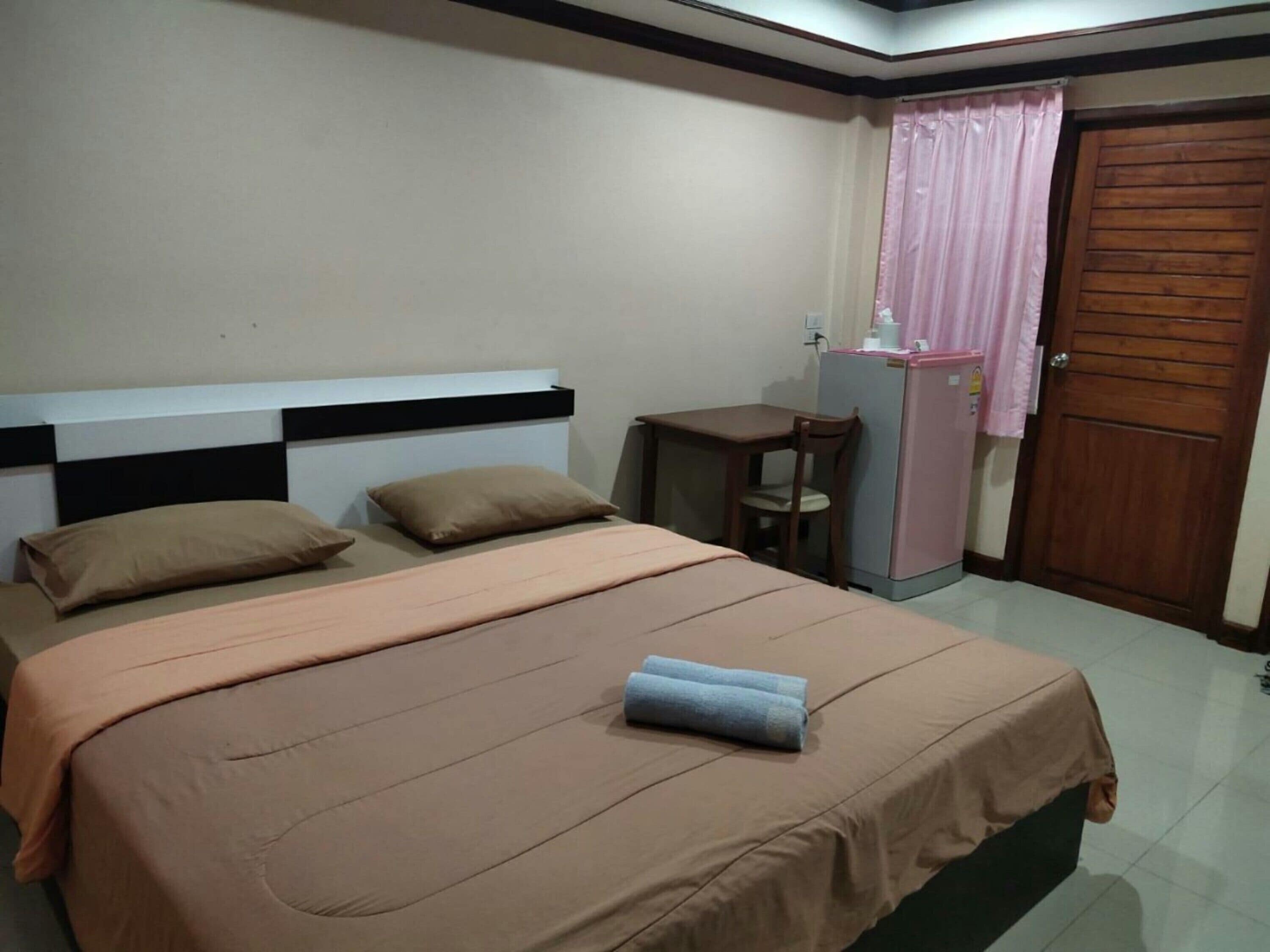 Un dormitorio con cama, dos almohadas, un escritorio con silla, un refrigerador y una puerta.