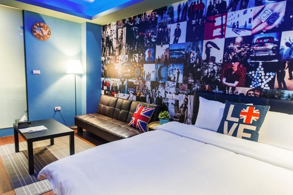 A hotel room with a large collage wall, a leather sofa, a bed with pillows, a bedside table, and a clock on the wall.