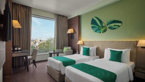 Quarto de hotel com duas camas, uma escrivaninha, uma cadeira, uma televisão e uma janela com vista para prédios.
