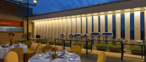 Un área de comedor al aire libre con mesas dispuestas para una comida, sillas y una barandilla con luces.