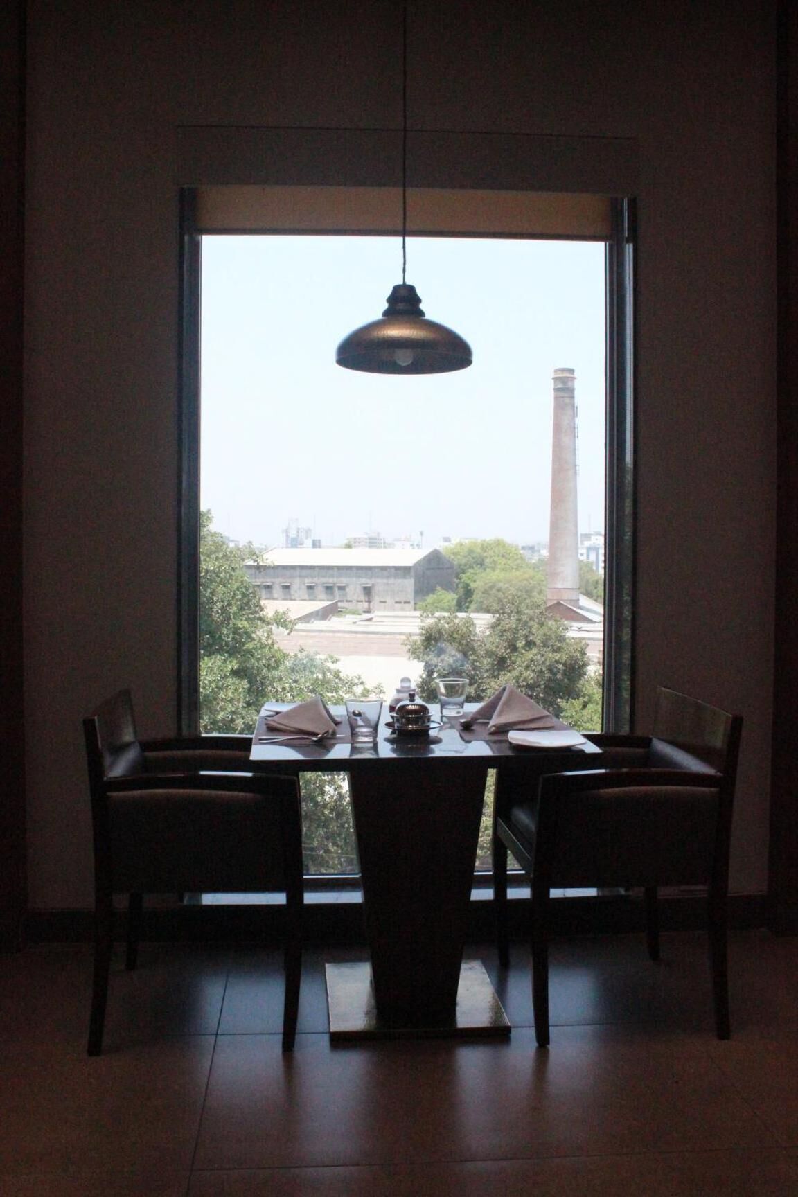 A dining area with a table set for two, a hanging light fixture, and a view of buildings and trees through a large window.