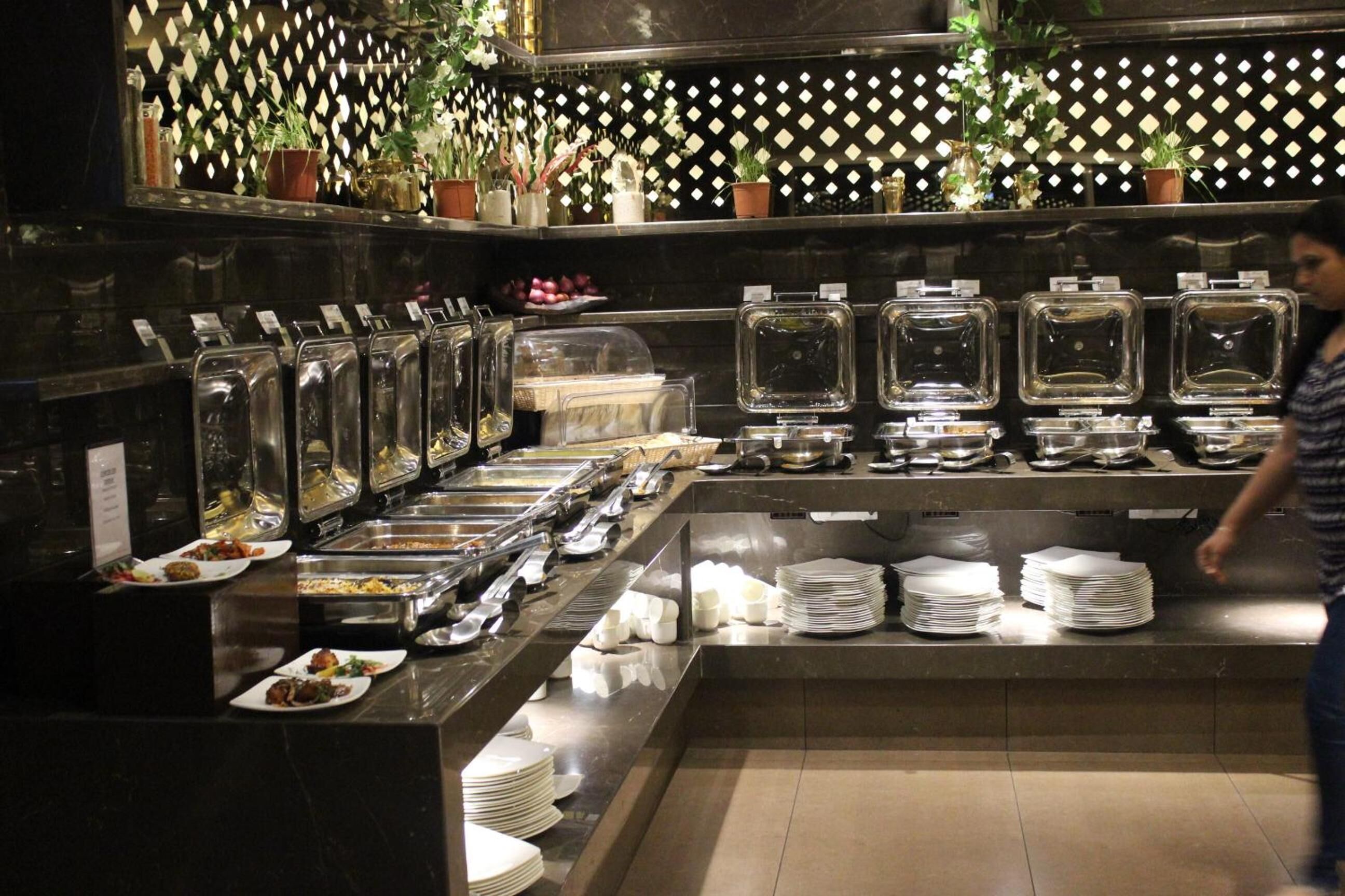 A buffet setup with various food containers and dishes, a person in the background, and a decorative wall with plants.
