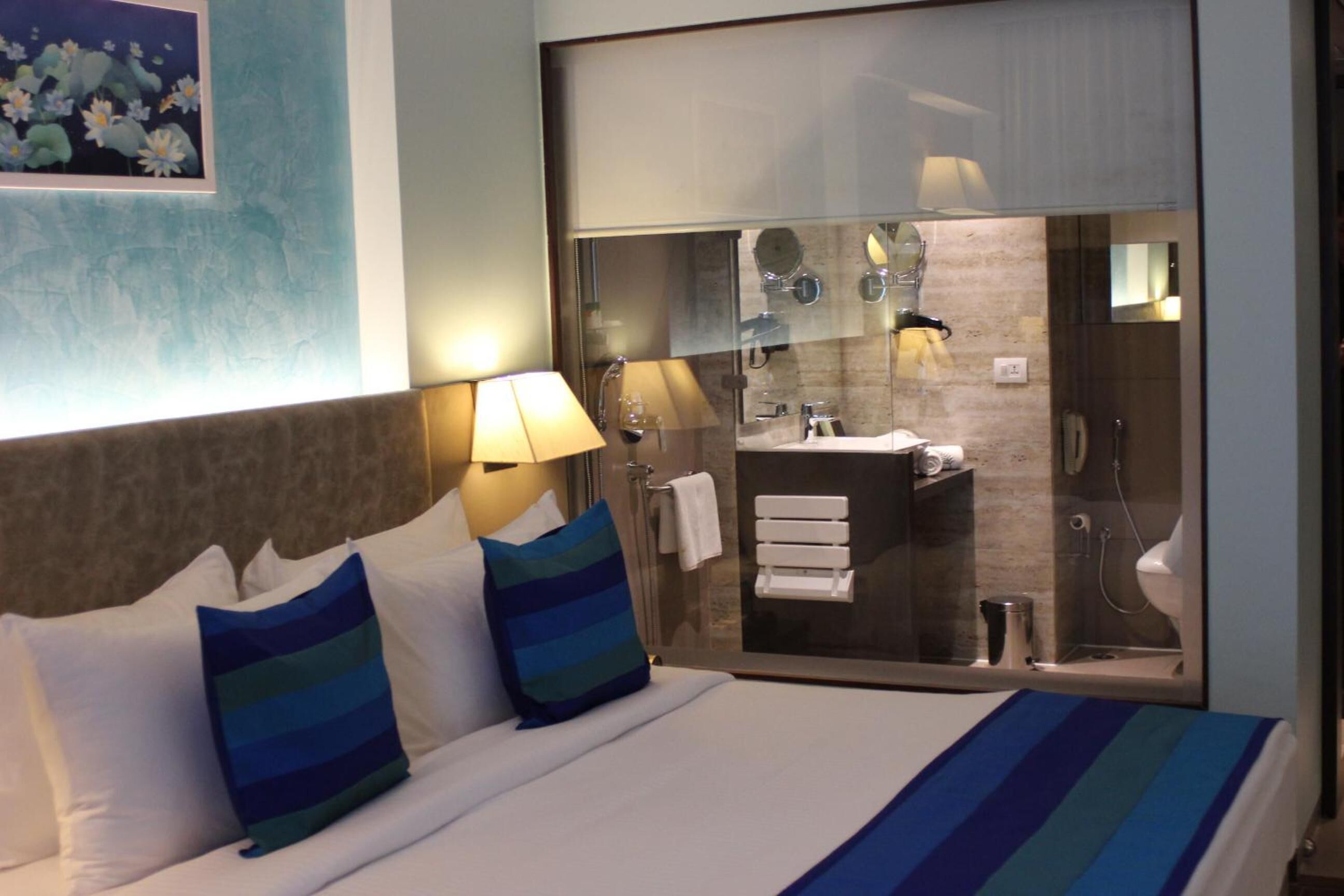 Habitación de hotel con cama, almohadas, mesita de noche, lámpara y vista al baño a través de una puerta de cristal.